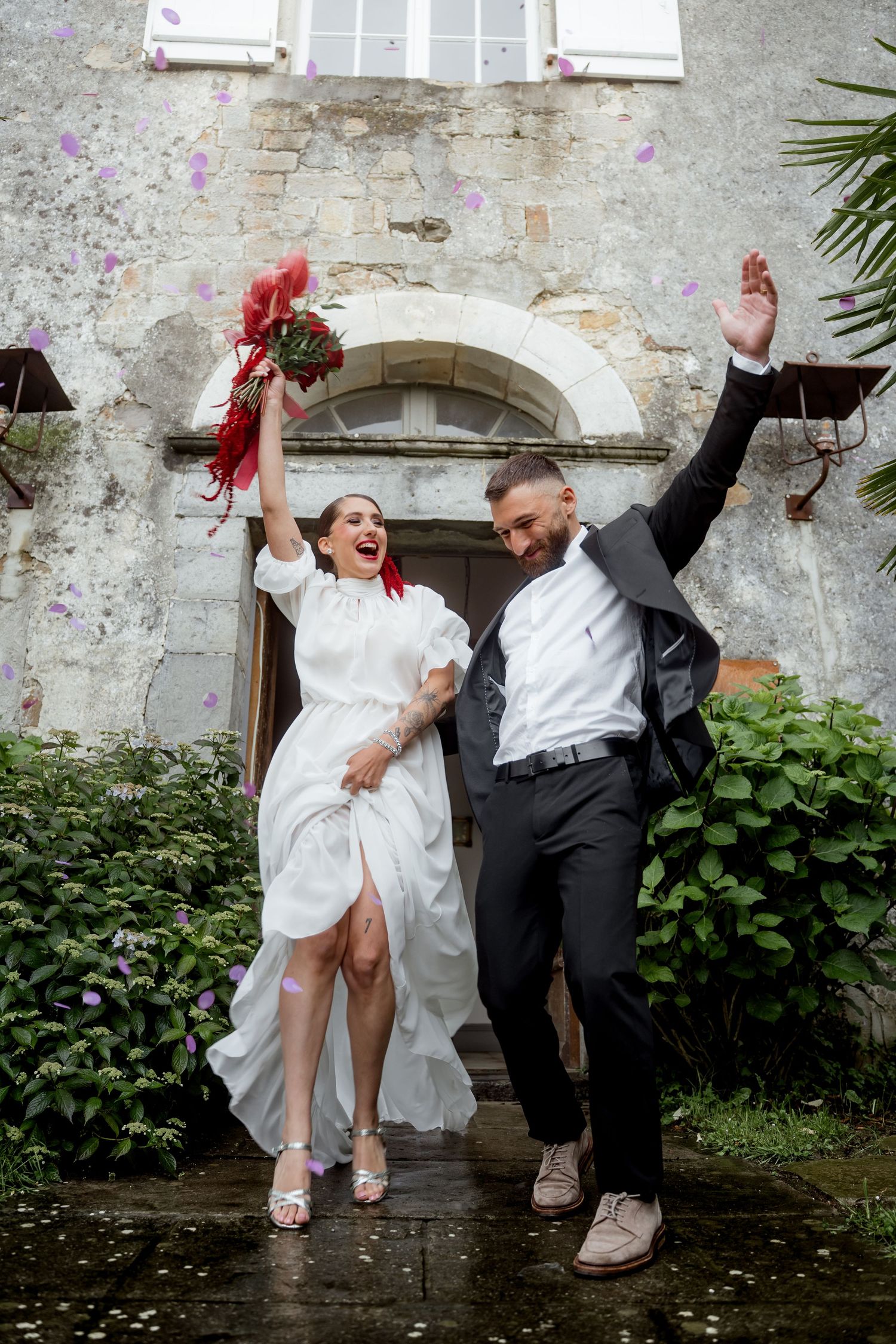 Excited bride and groom celebrate in rain outside rustic stone venue with red bouquet.