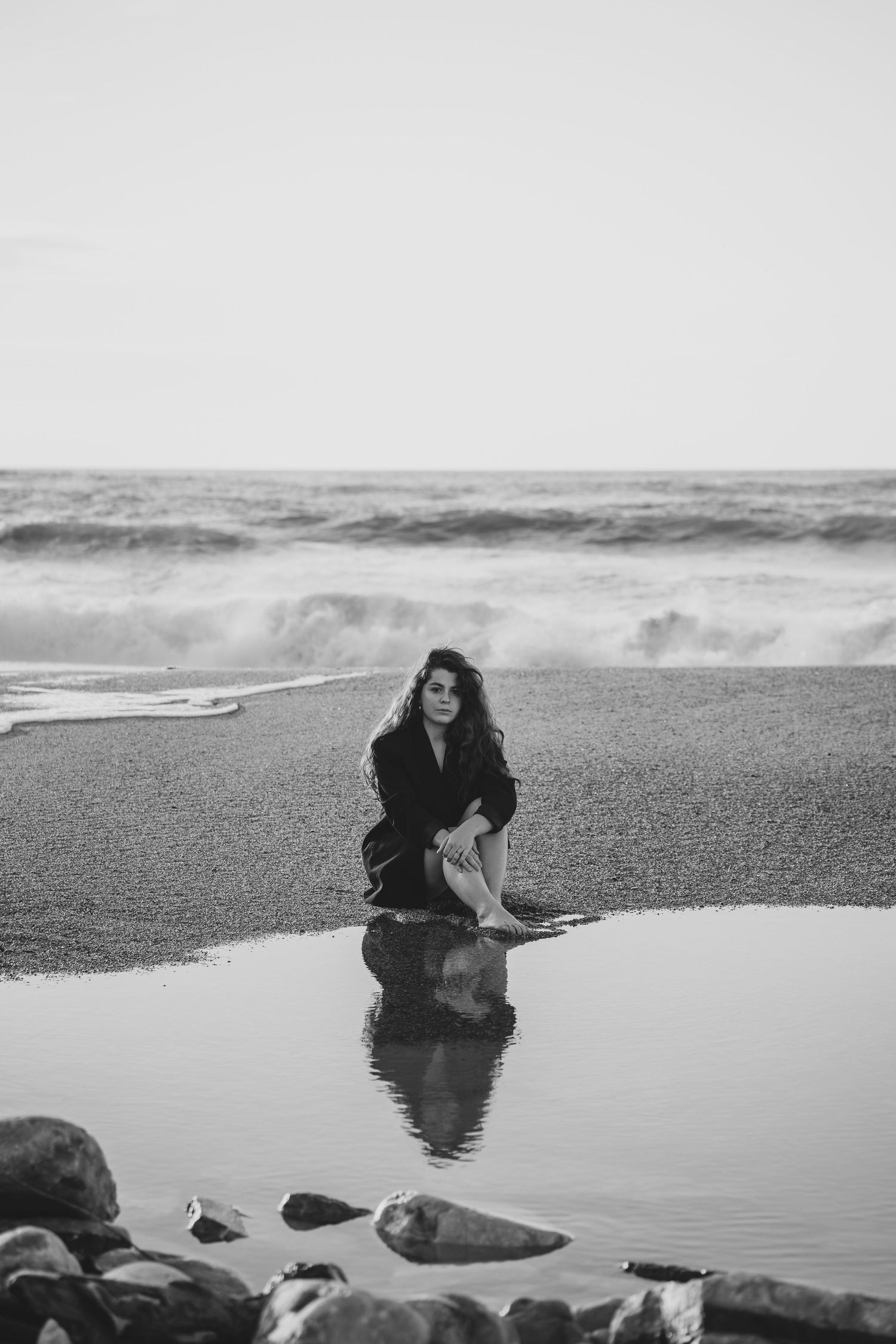 Black and white photo of a woman in a dark outfit kneeling on a beach with her reflection in a tide pool and waves behind her.