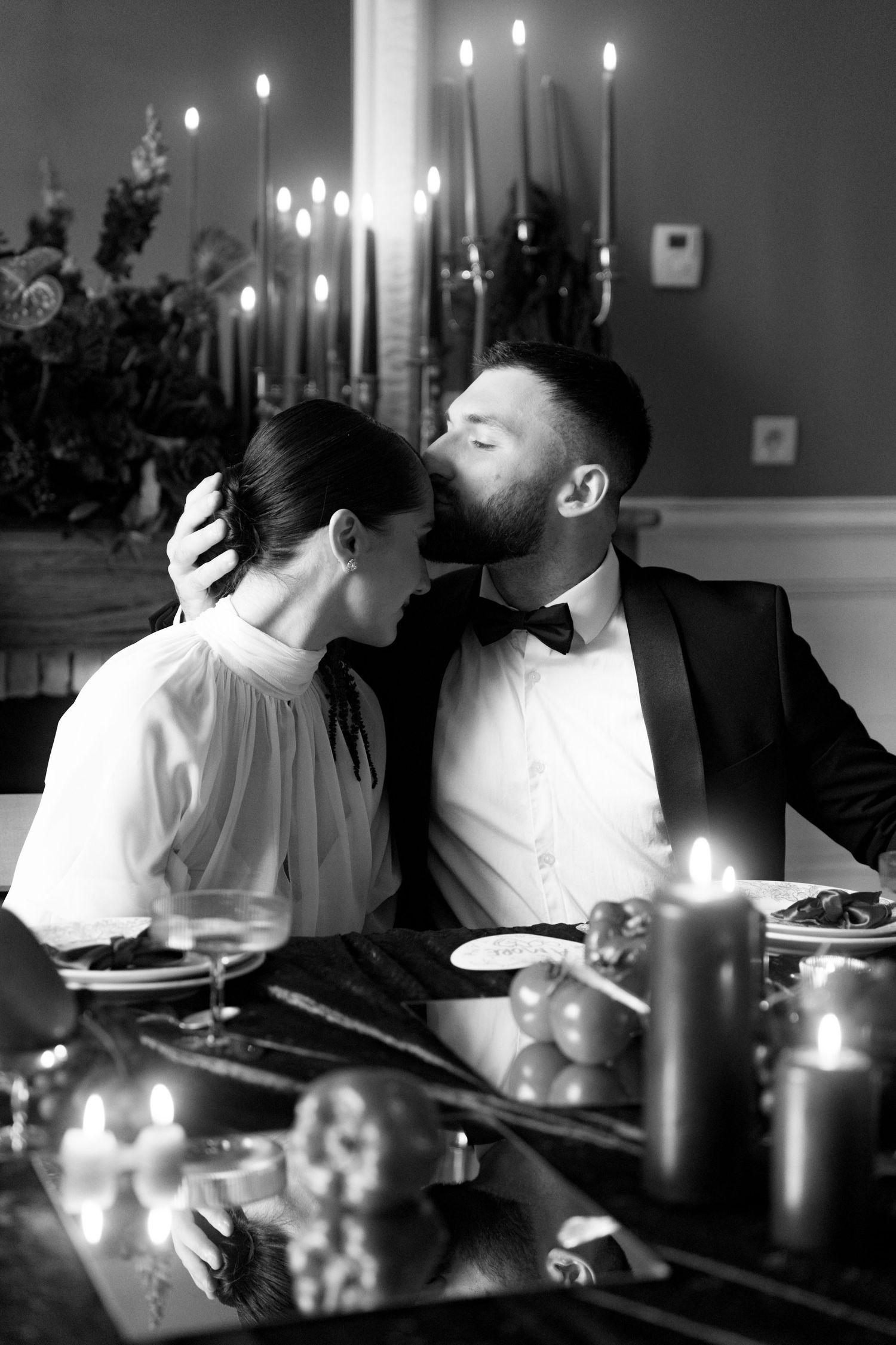 Black and white photo of bearded groom in tuxedo kissing bride at romantic candlelit dinner.