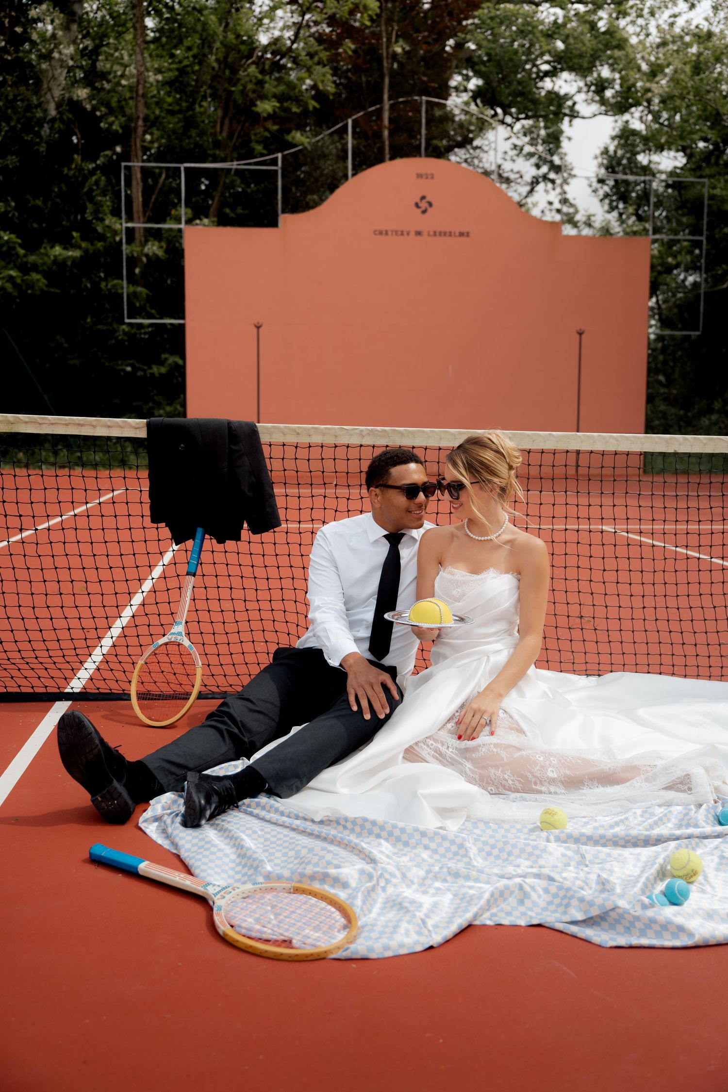 Bride and groom relaxing on red clay tennis court surrounded by rackets and tennis balls.