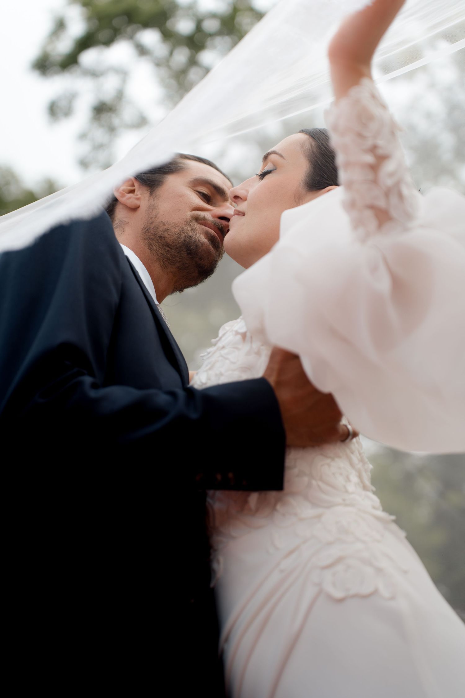Bride and groom sharing a romantic kiss under a flowing white veil outdoors.