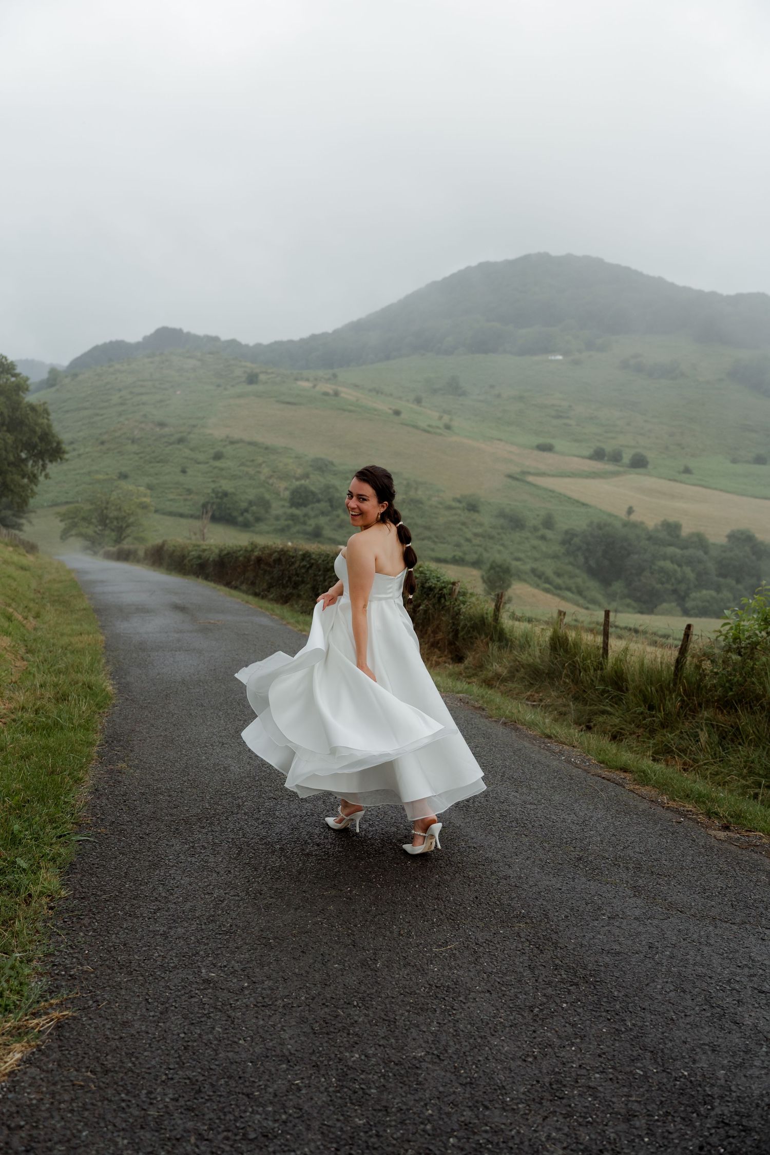Bride in white wedding dress twirling on a misty country road surrounded by green hills.