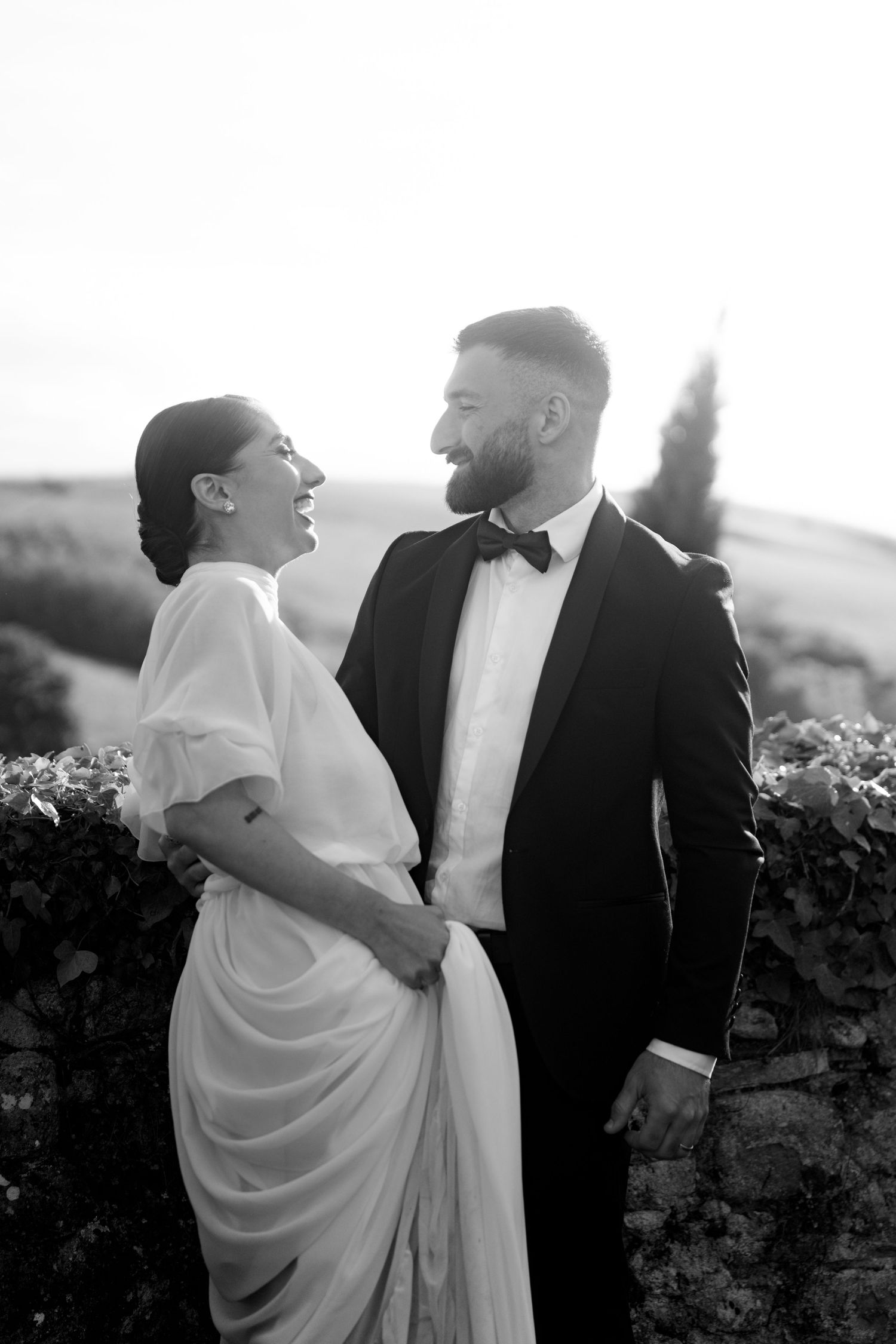 Black and white photo of laughing bride and bearded groom sharing joyful moment in scenic countryside.