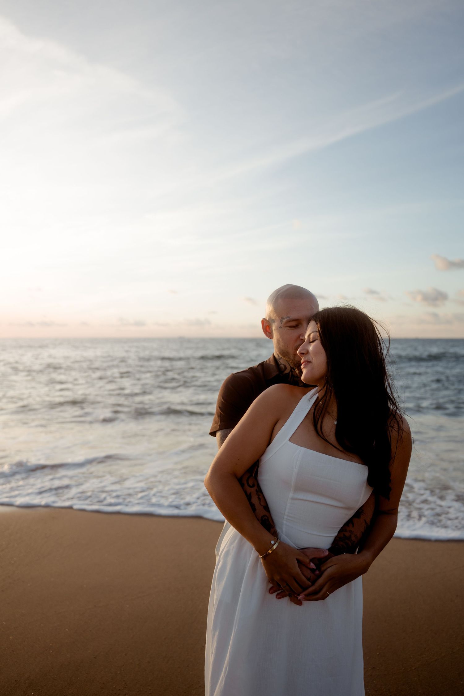 Bald tattooed man embracing woman in white dress from behind on a beach during a pastel sunset.