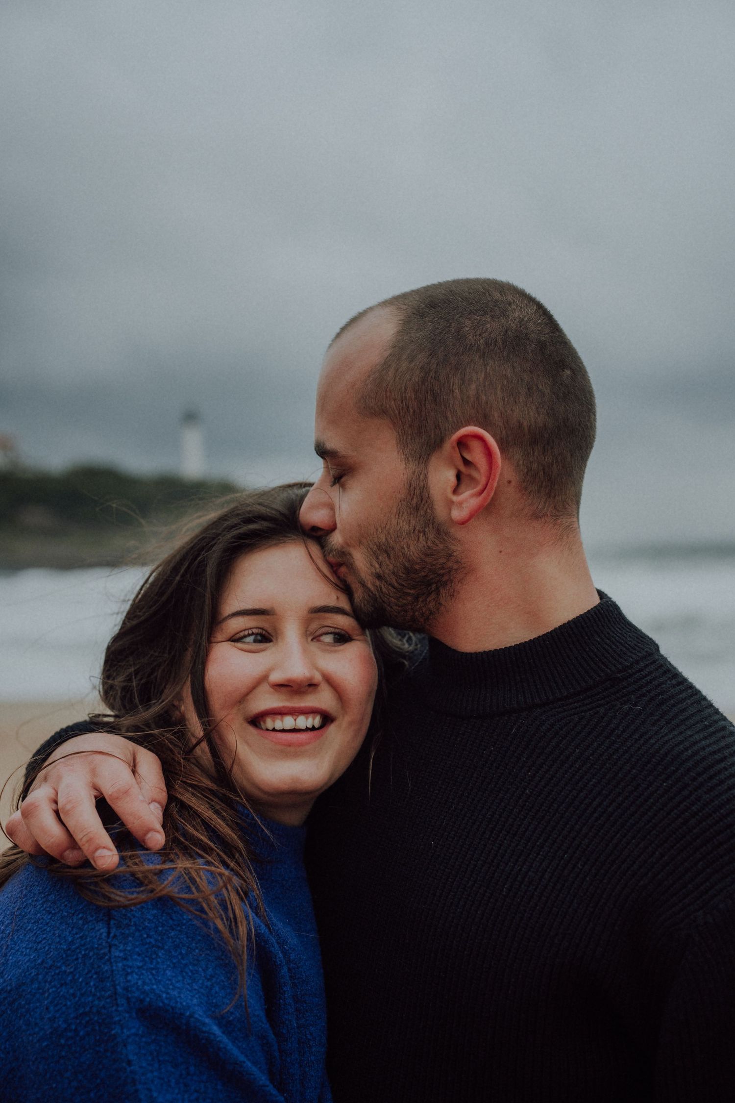 Man kissing smiling woman on the head at a beach, woman in blue sweater laughing joyfully outdoors.