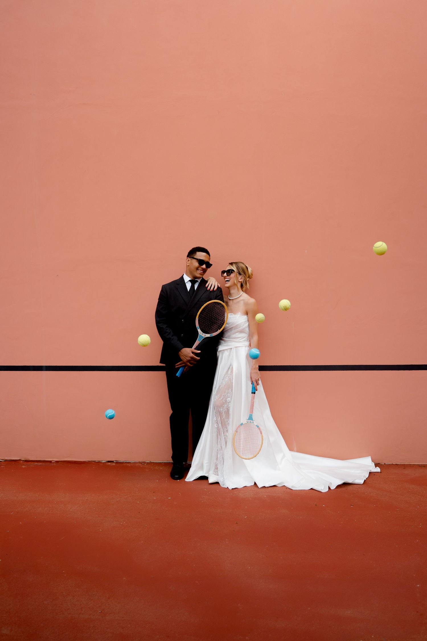 Bride and groom in sunglasses pose against pink tennis court wall with colorful balls flying.