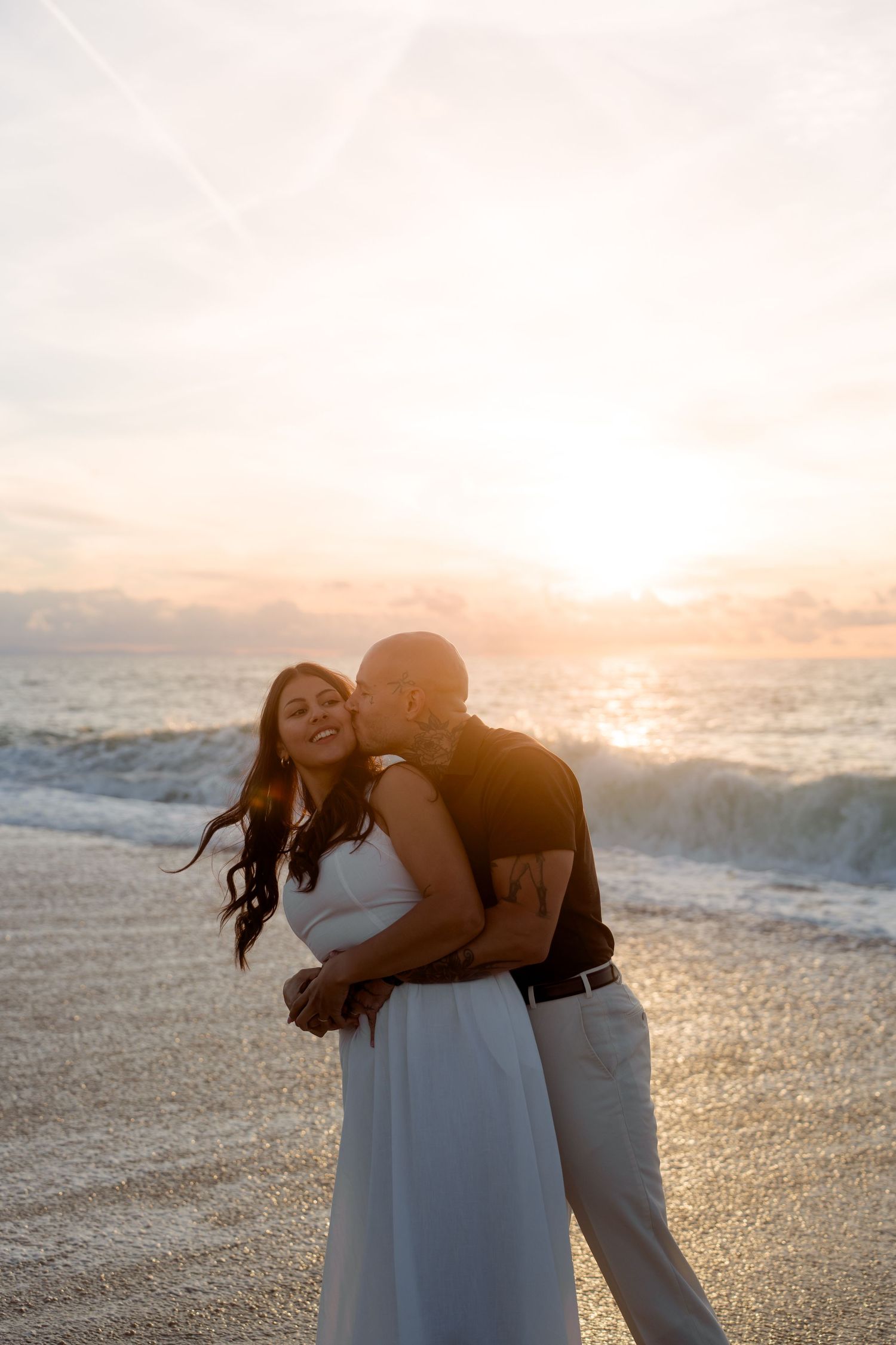 Couple sharing a romantic kiss on a beach during a golden sunset with ocean waves behind them.