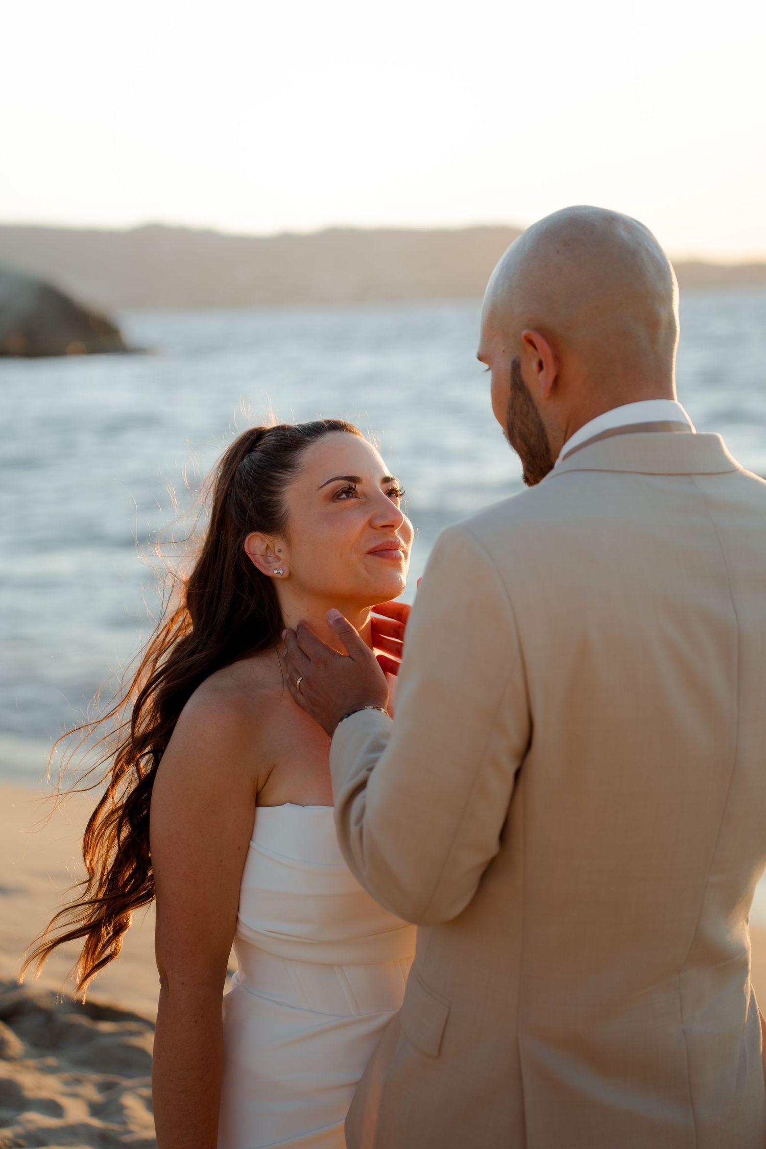 Groom gently cupping brides chin as she smiles up at him on a sunlit beach.