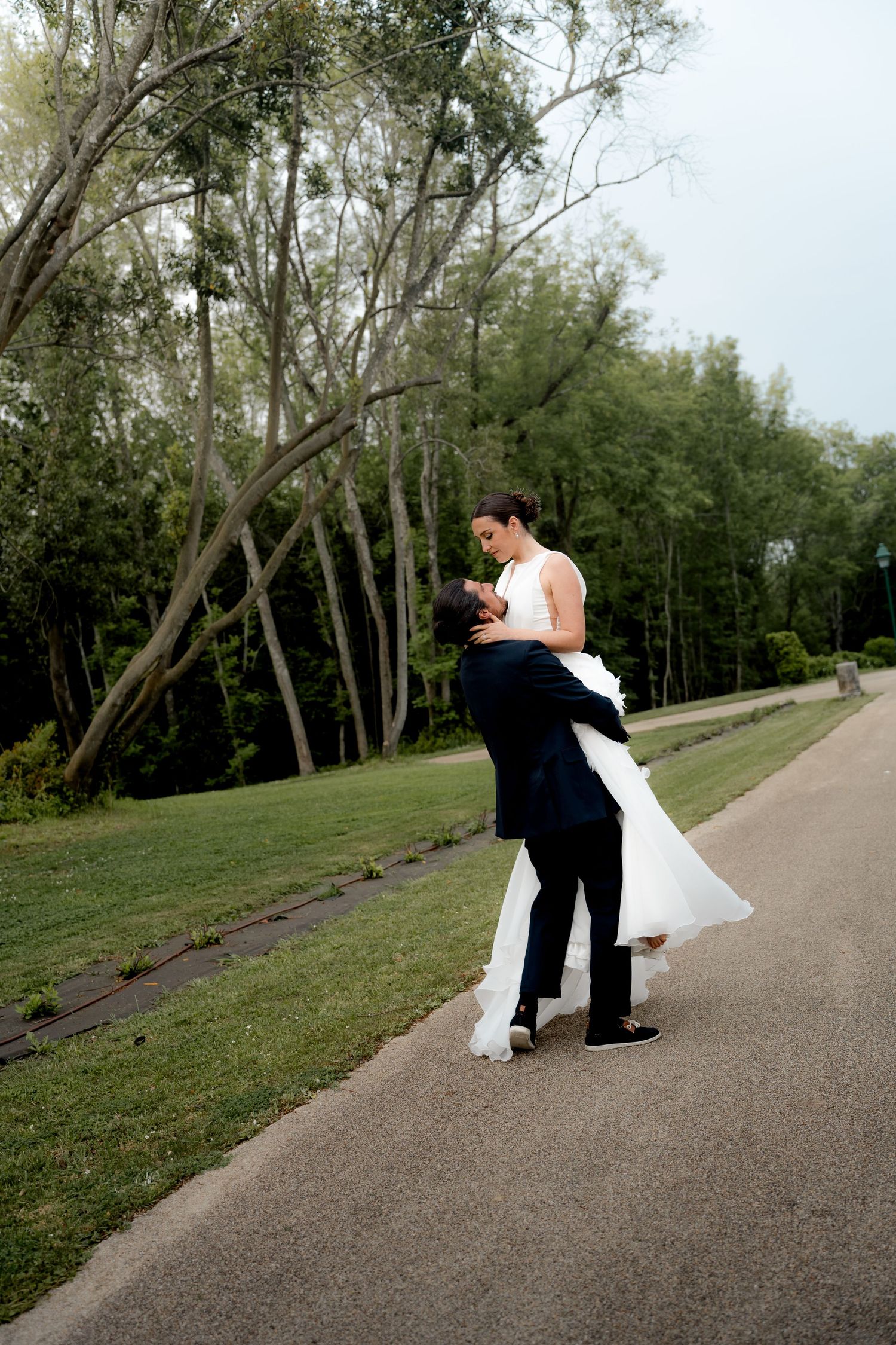 Groom lifts bride in white wedding gown on a tree-lined pathway in a lush green park.