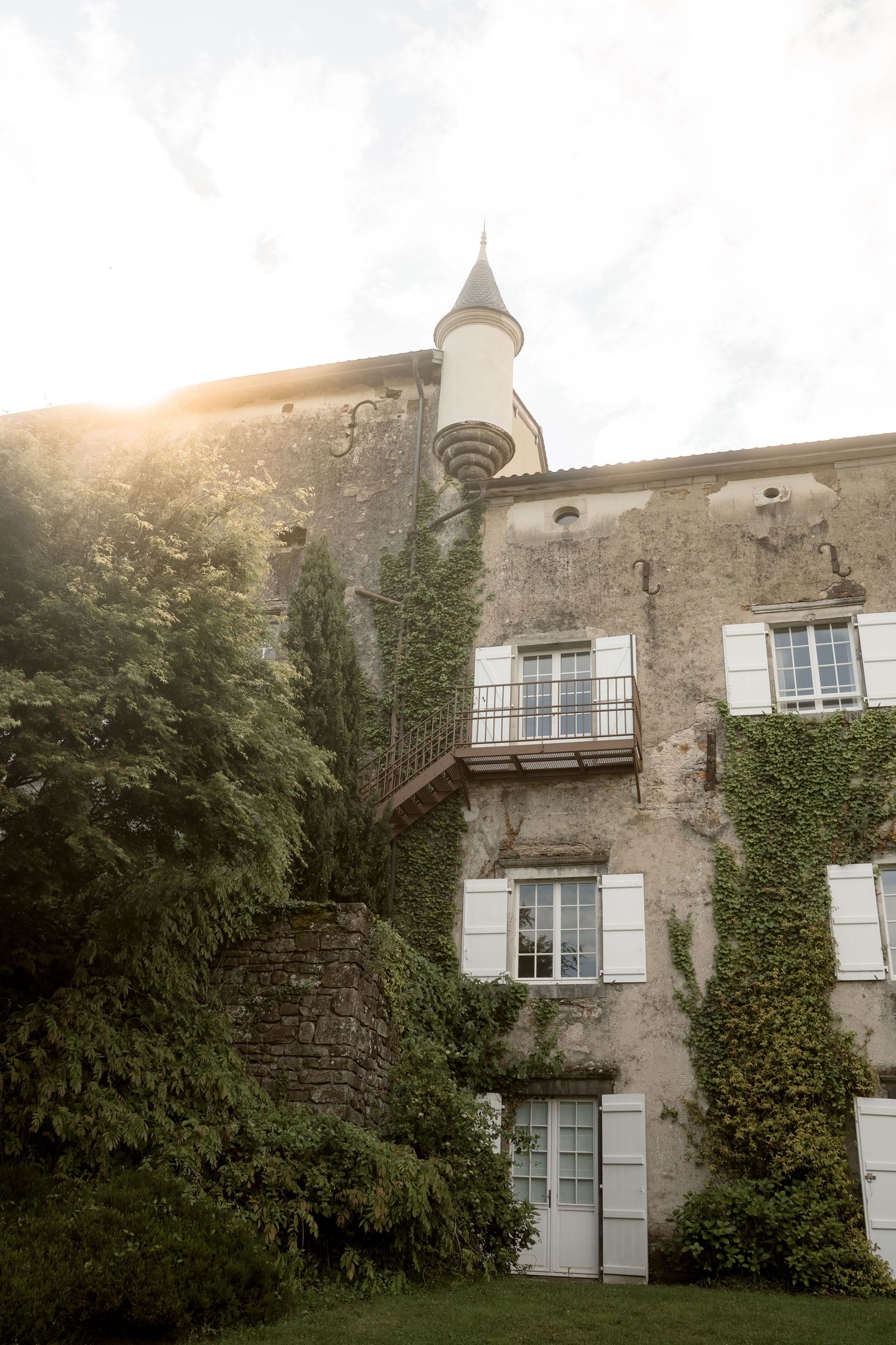 Ivy-covered French château with white shuttered windows, turret, and lush greenery bathed in golden sunlight.