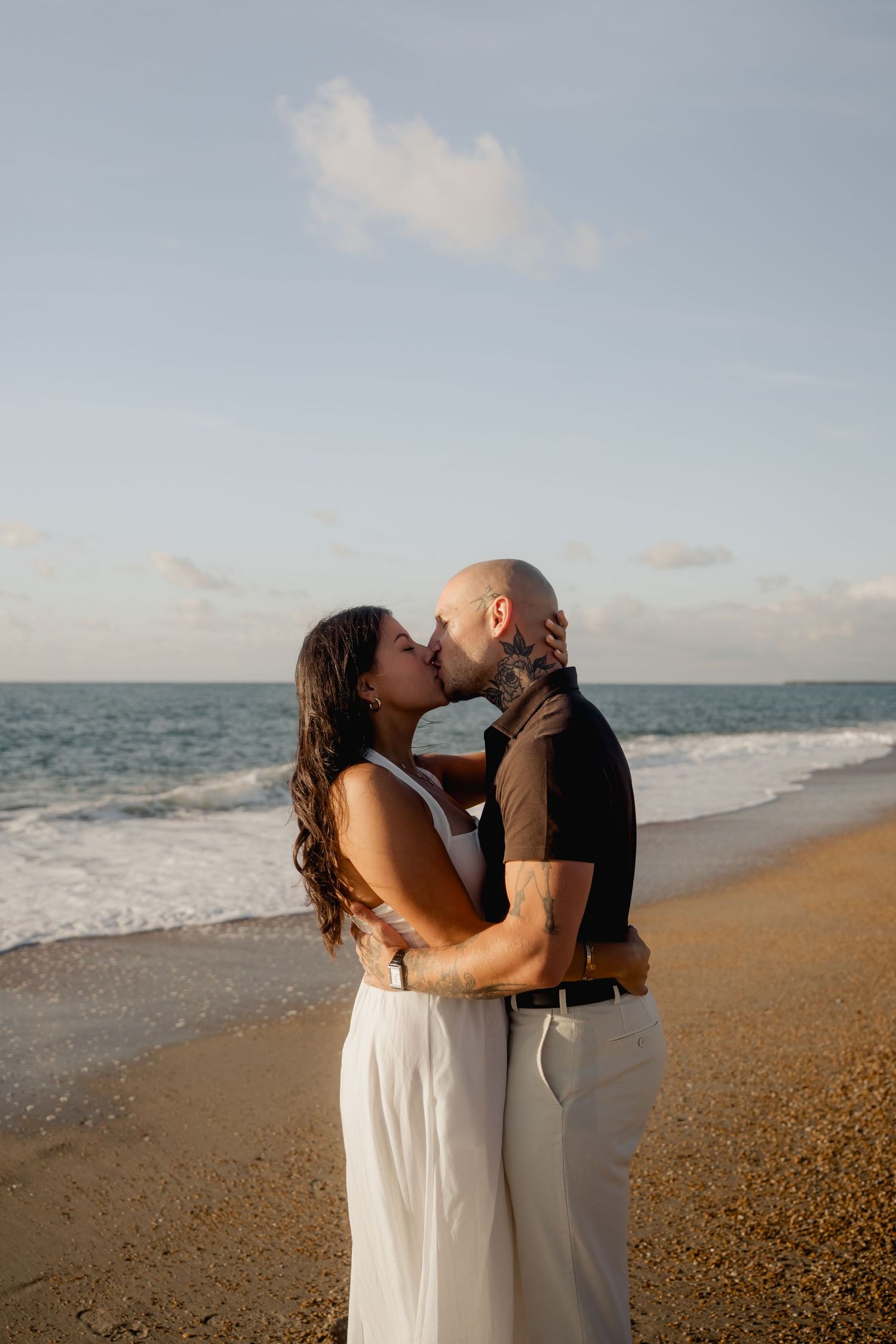 Couple sharing romantic kiss on beach at sunset, bald tattooed man embracing woman in white dress.