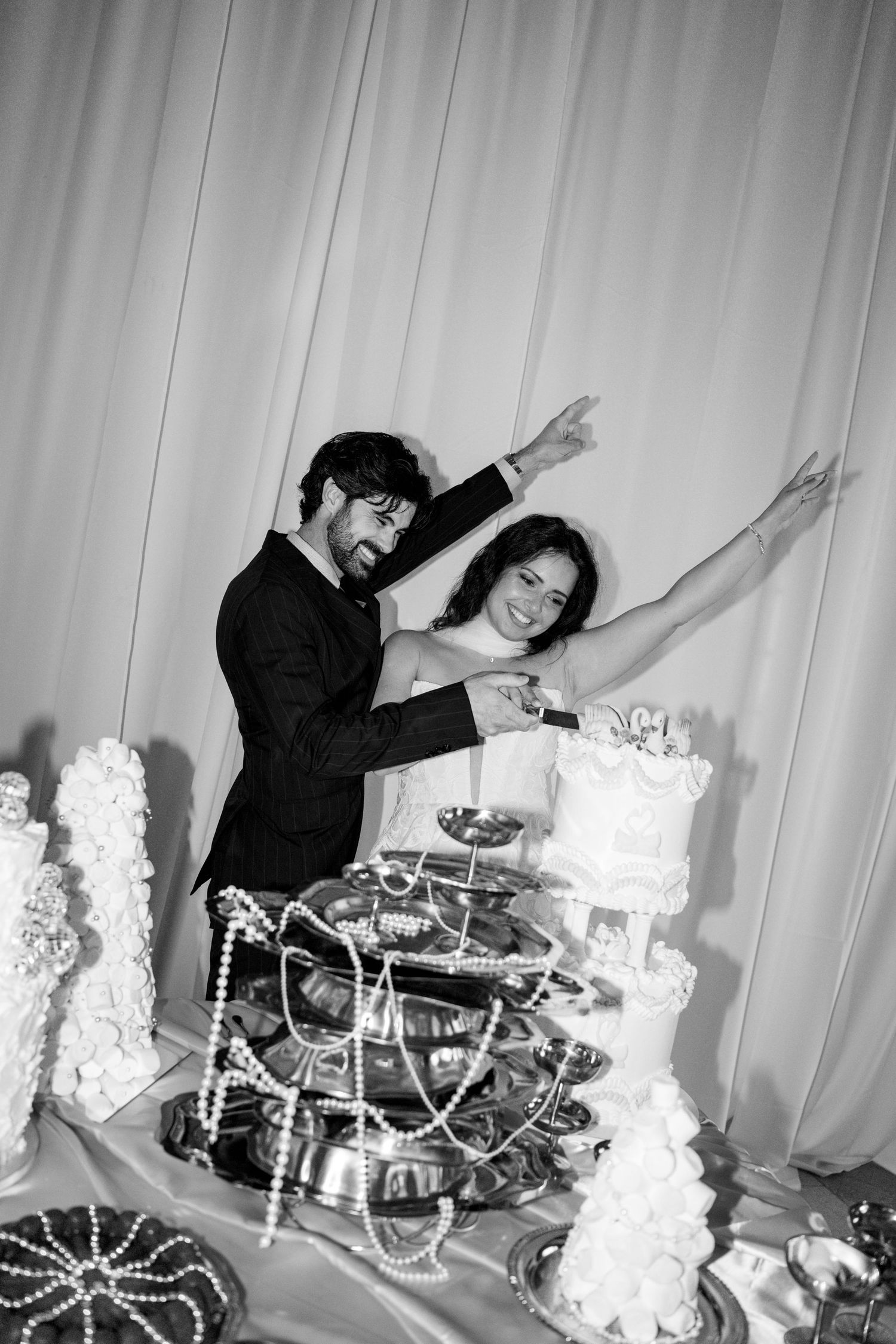 Black and white photo of joyful bride and groom cutting their wedding cake with arms raised in celebration.