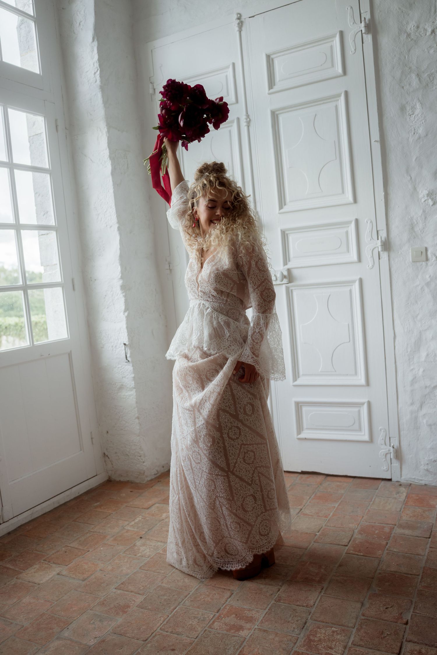 Bohemian bride in lace bell-sleeve gown raising red bouquet overhead in whitewashed rustic room.