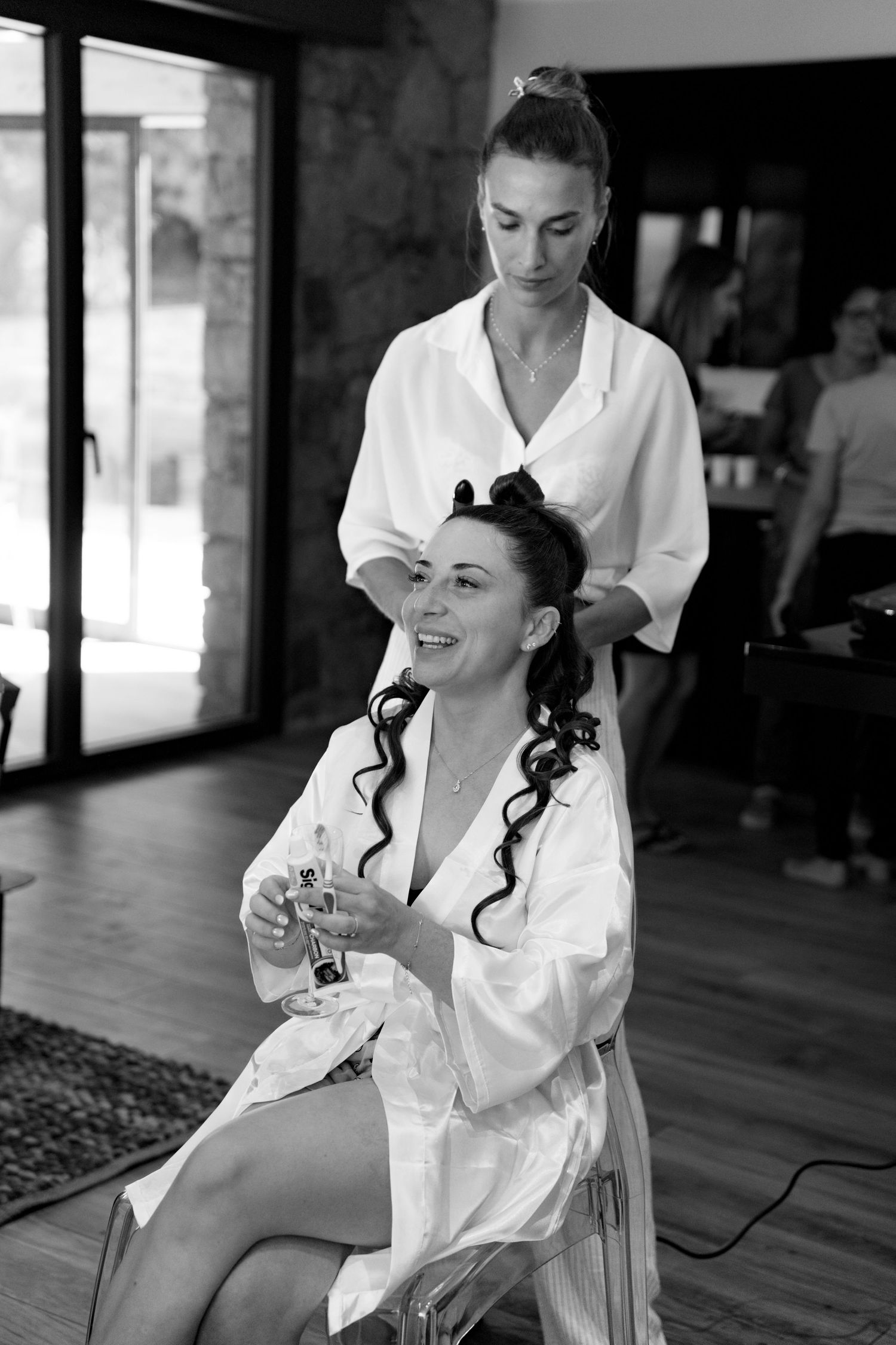 Black and white photo of bride laughing while hairstylist styles her curly hair during wedding preparations.