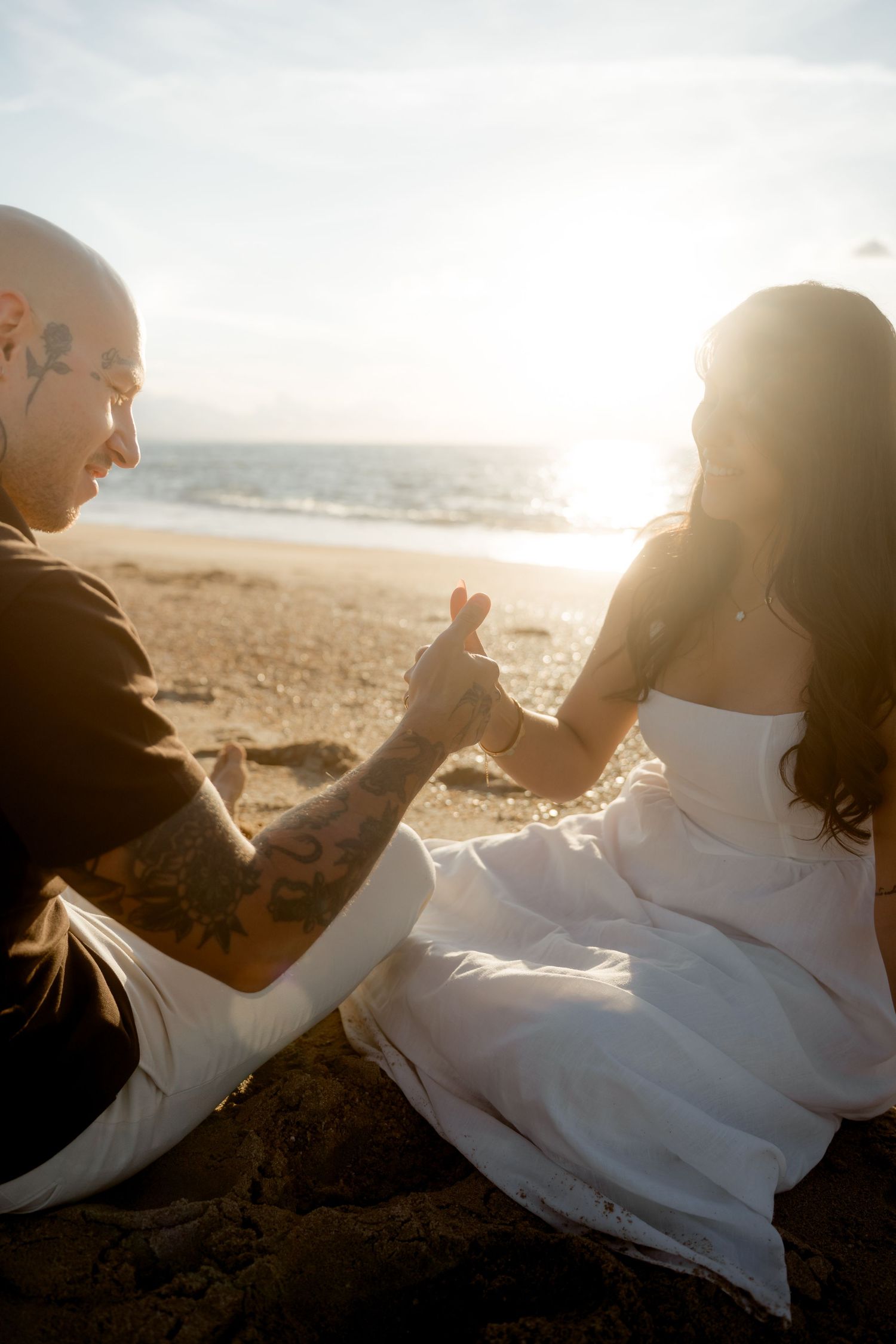 Tattooed man and woman sitting on beach holding hands in warm golden sunlight.