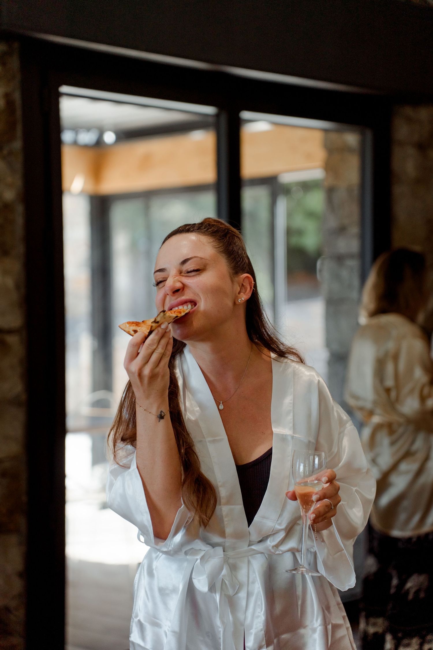 Joyful bride in white satin robe eating pizza and holding champagne glass during wedding morning preparations.