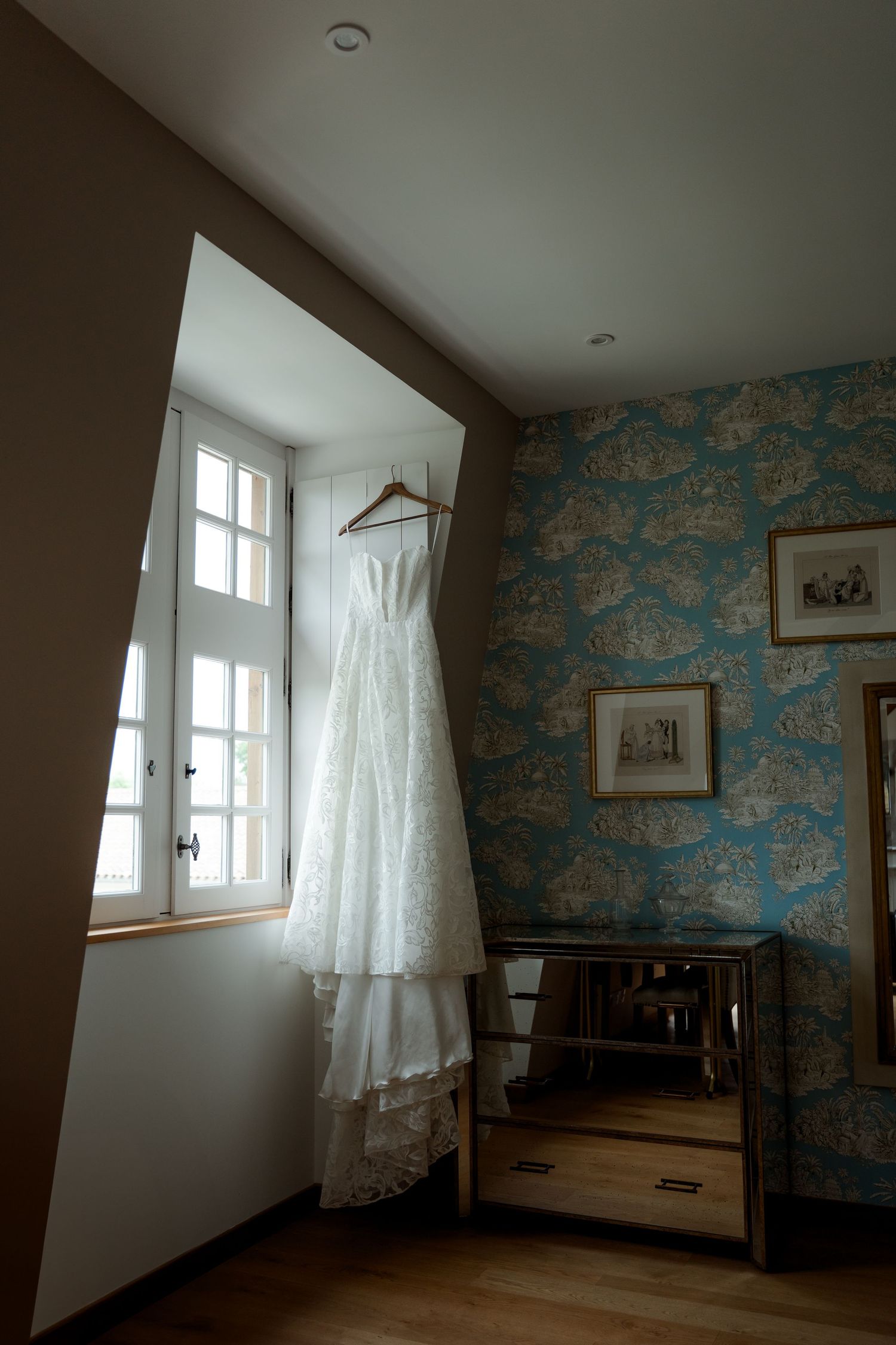 White lace wedding dress hanging on wooden hanger in window of room with blue toile wallpaper.