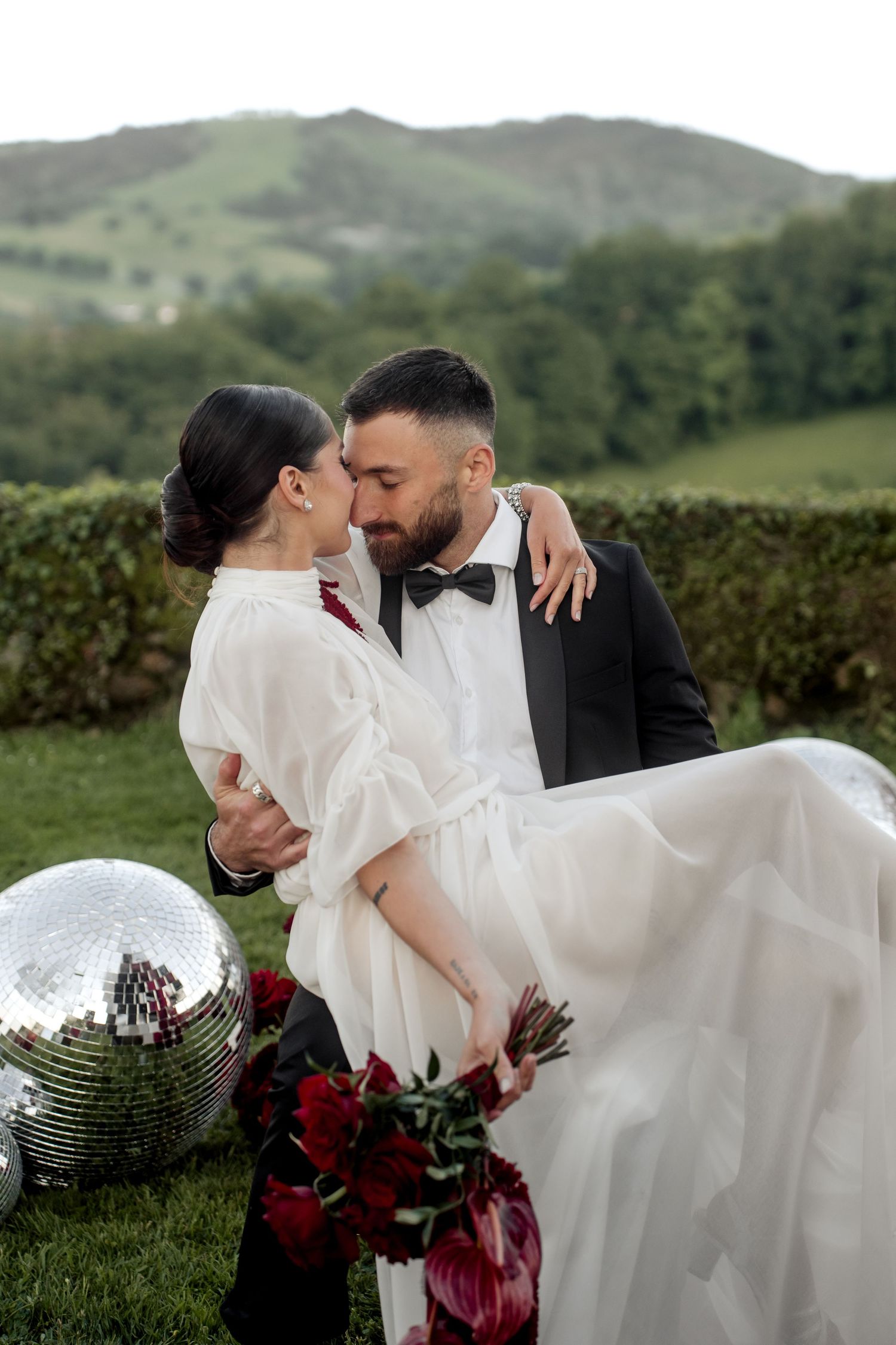 Groom lifts bride in white ruffled dress near disco balls with lush green hills in the background.