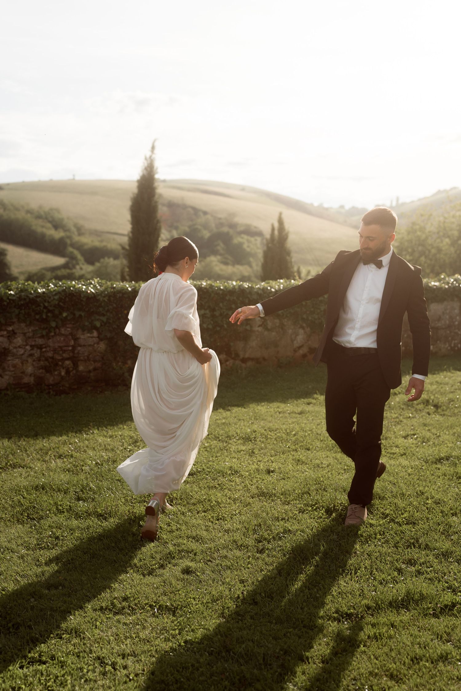Bride and groom playfully running on green lawn with rolling Tuscan hills in background.