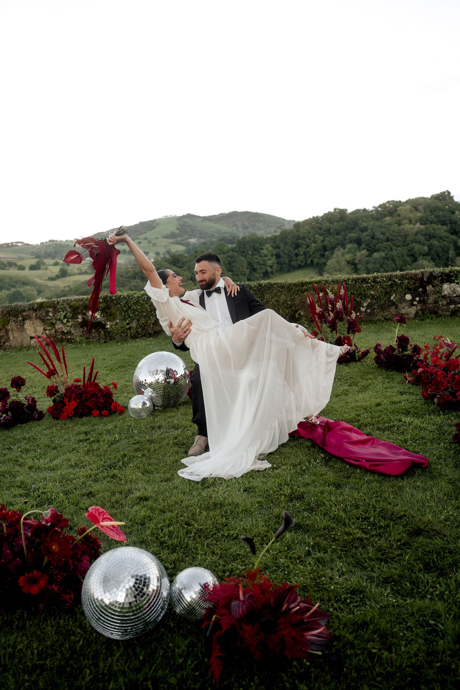 Groom lifts bride in flowing white dress surrounded by red florals and disco balls on green lawn.