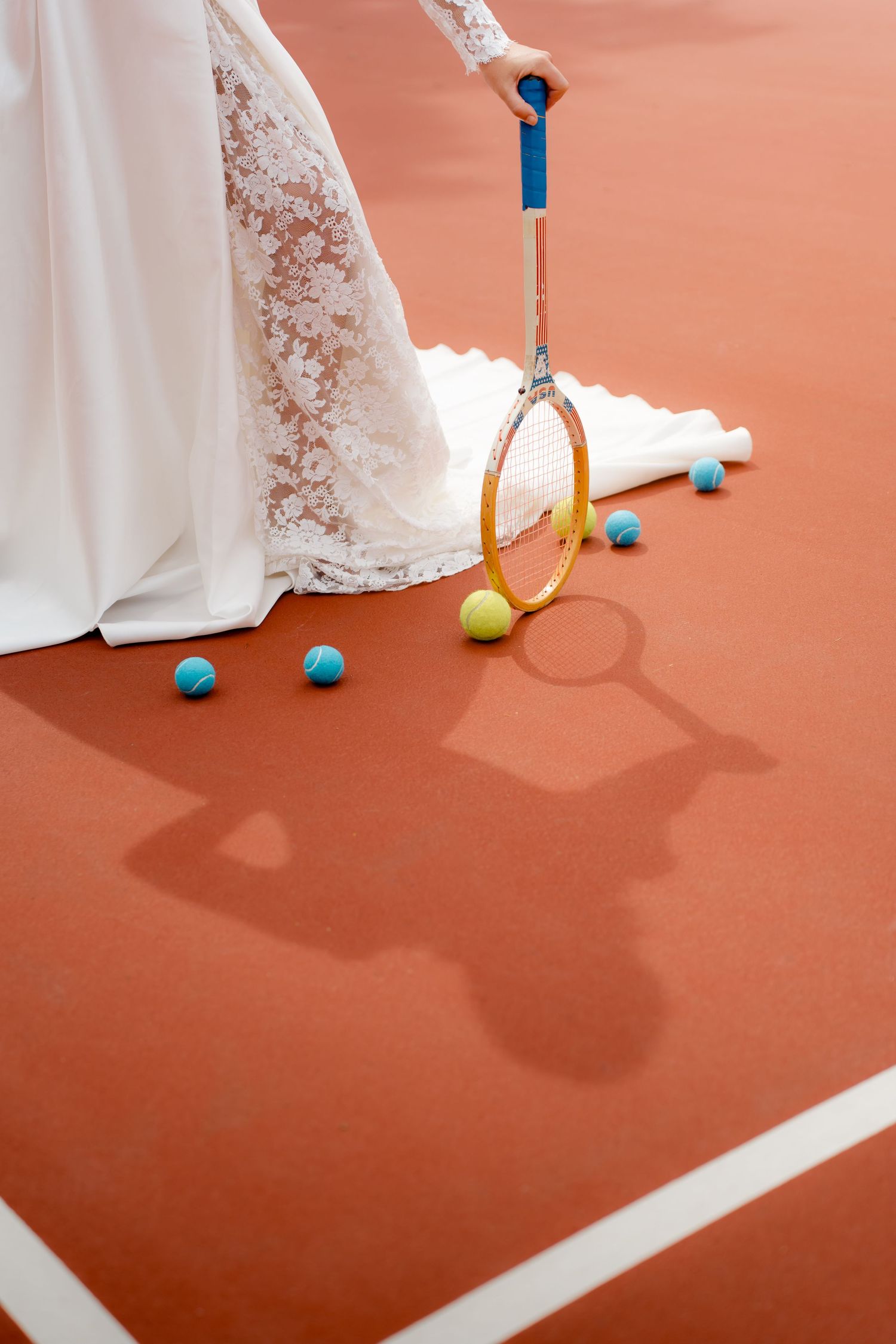 Close-up of bride's white lace gown with vintage tennis racket and colorful balls on clay court.