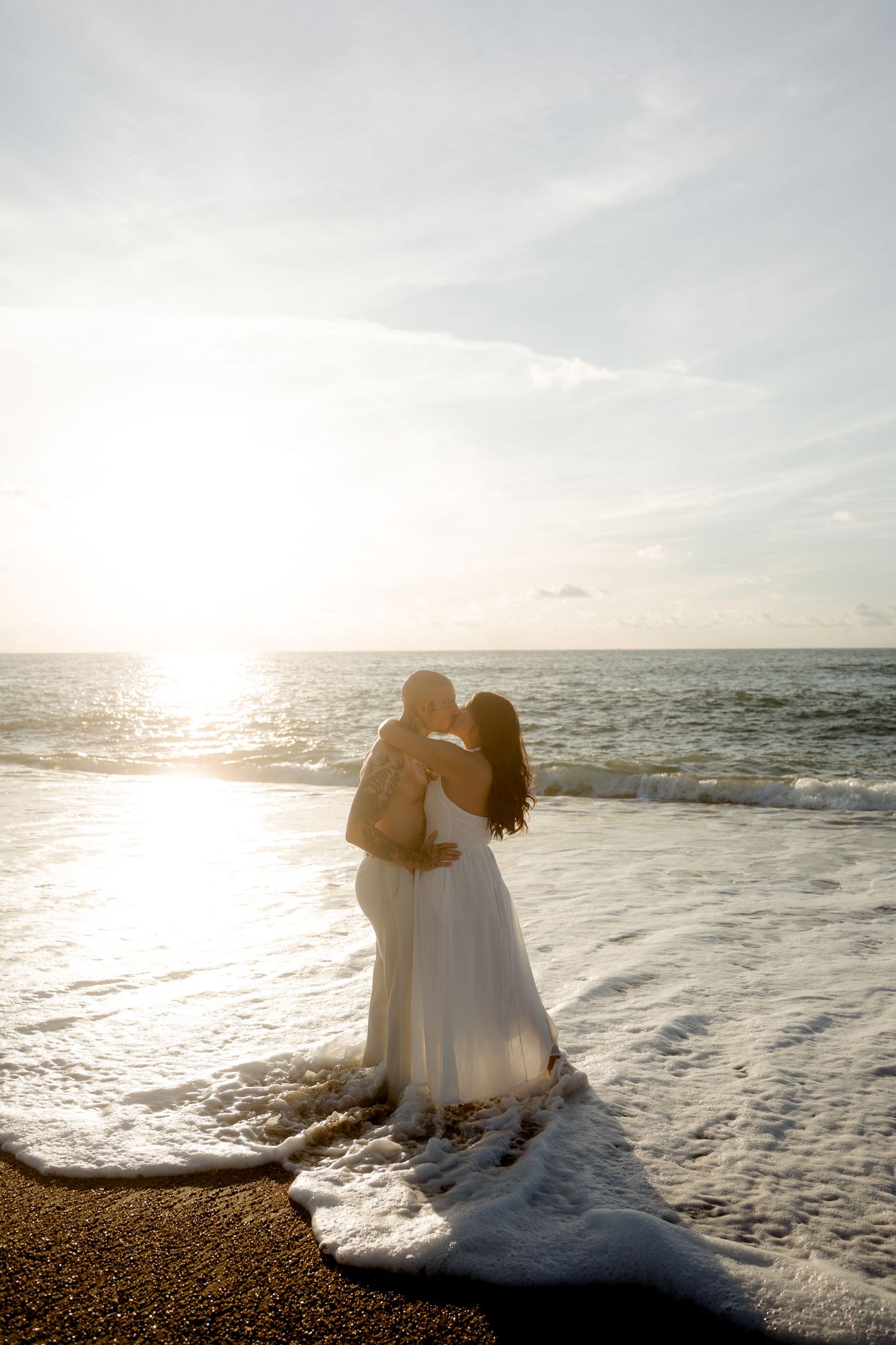 Couple embracing and kissing on beach at sunset, ocean waves surrounding woman's white dress.