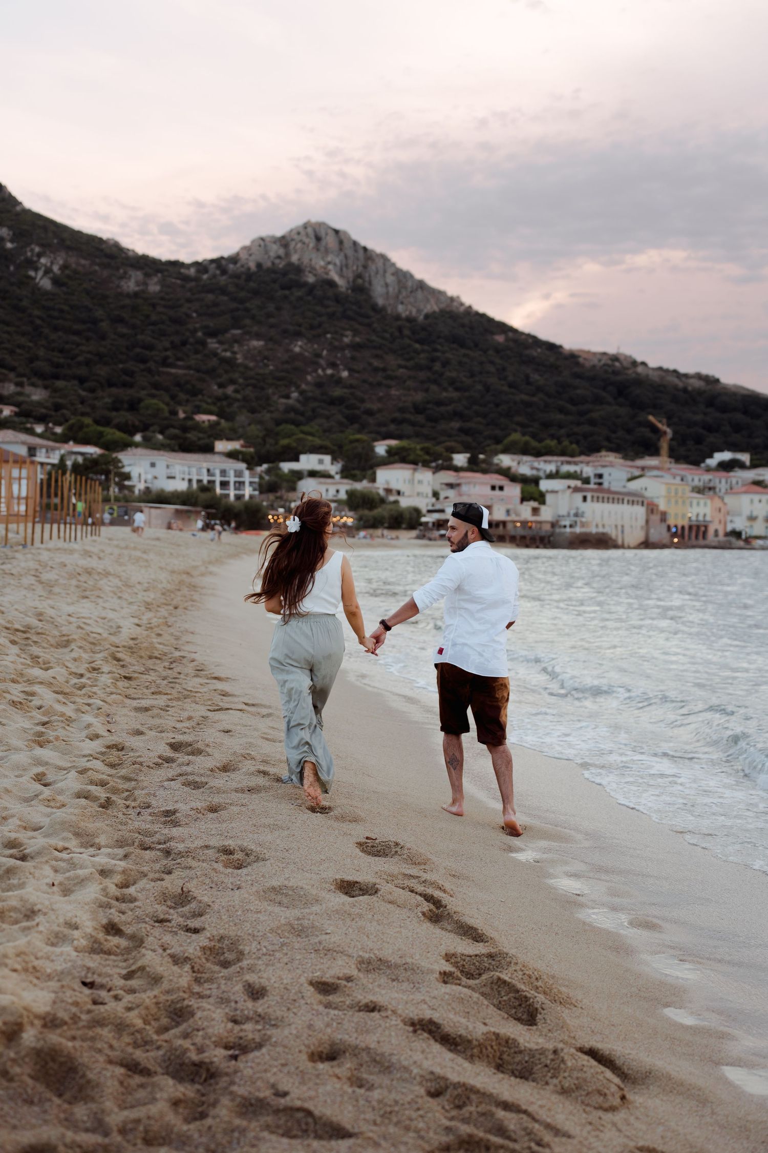 Couple holding hands running along a sandy beach at sunset with a mountain backdrop.
