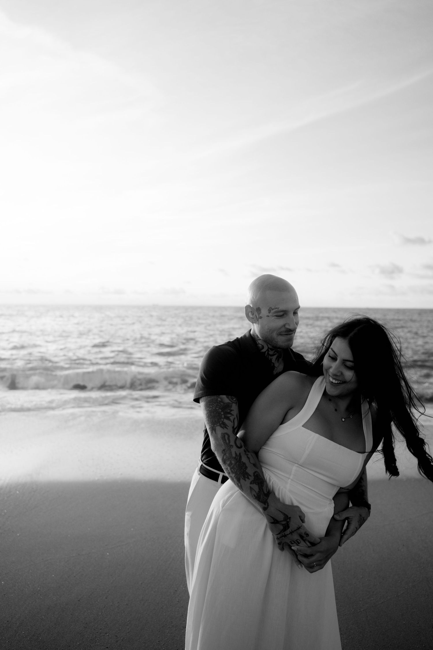Black and white photo of tattooed bald man embracing smiling woman in white dress on a beach.