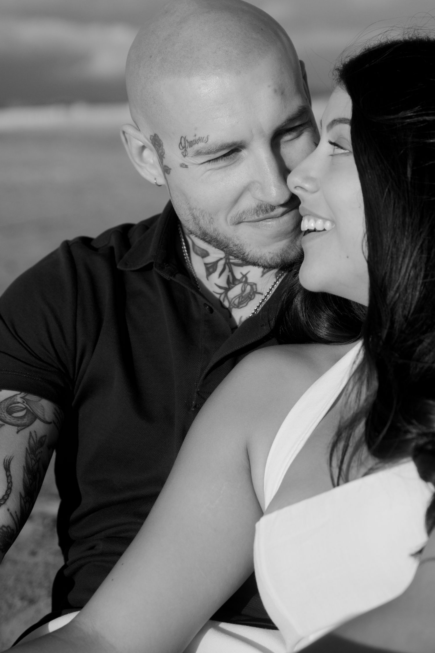 Black and white photo of tattooed bald man smiling as woman kisses his cheek on beach.