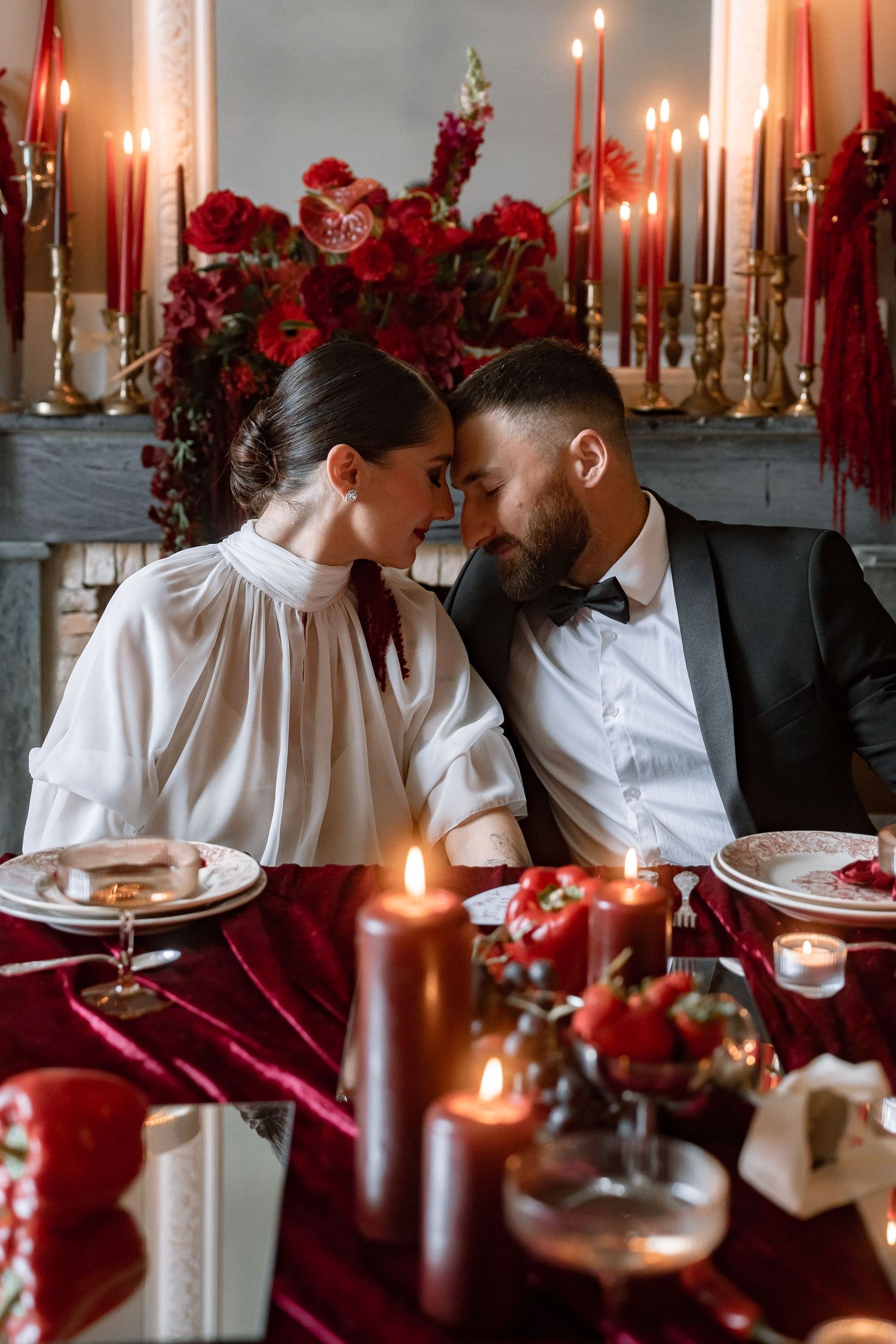 Couple shares romantic moment at candlelit wedding table surrounded by red roses and candles.