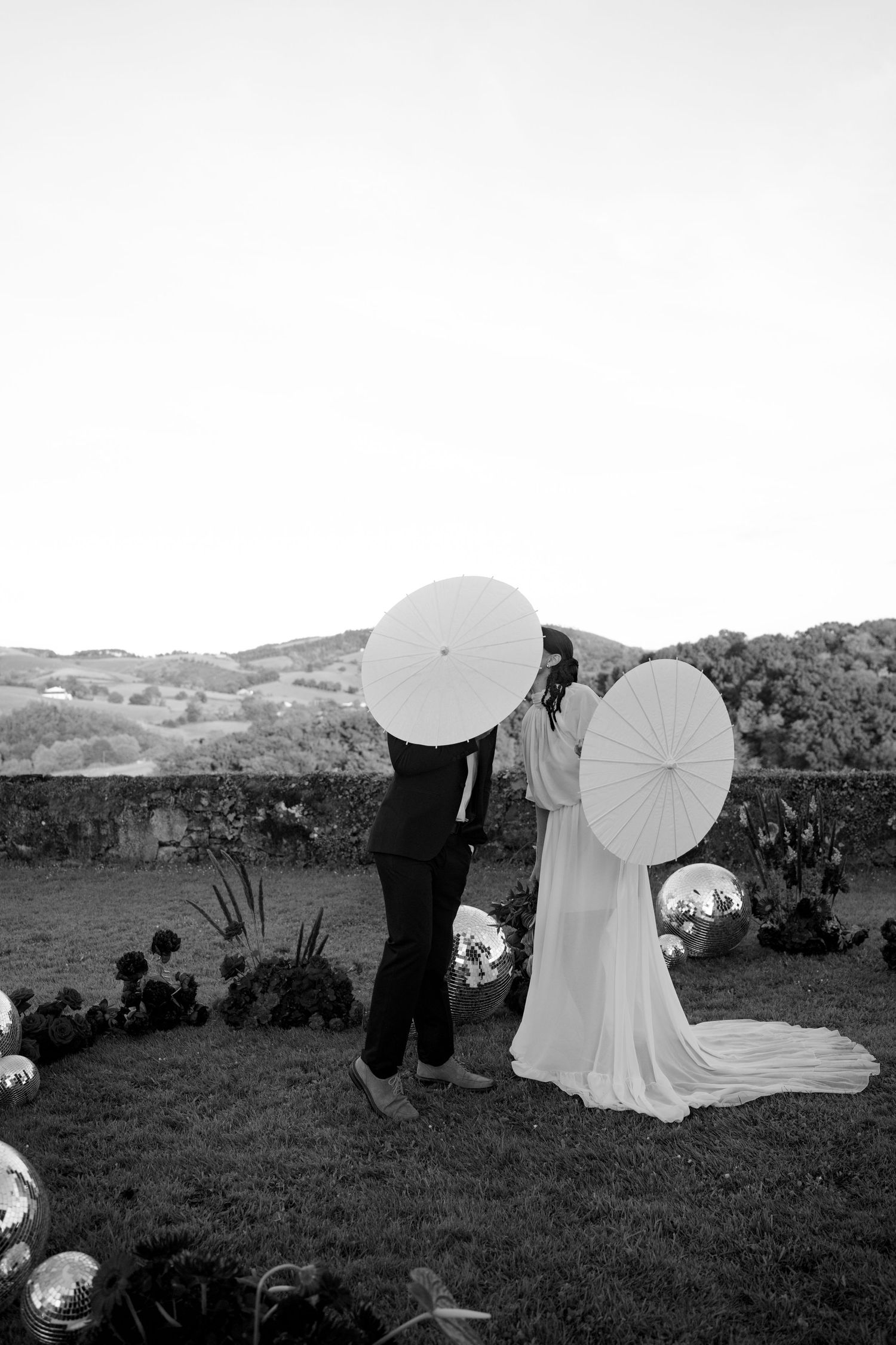 Black and white photo of bride and groom hiding behind white parasols at their outdoor wedding.