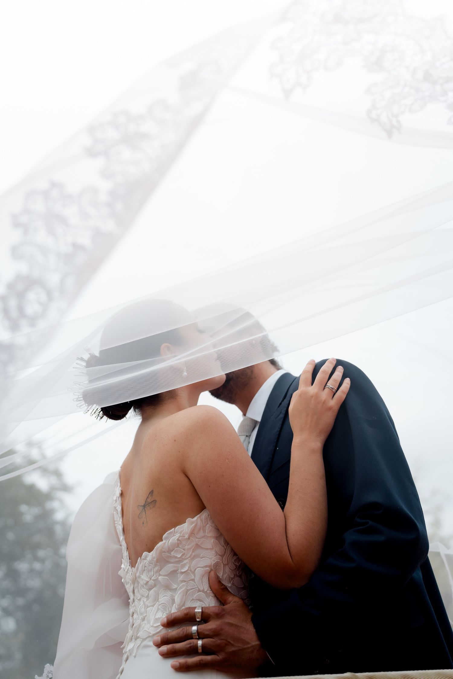 Bride and groom kissing beneath billowing lace-trimmed veil with arms around each other.