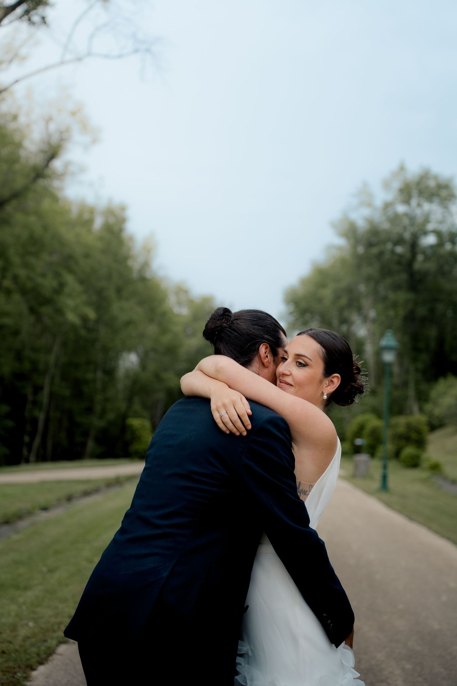 Bride in white gown embraces groom on scenic park pathway surrounded by lush green trees.