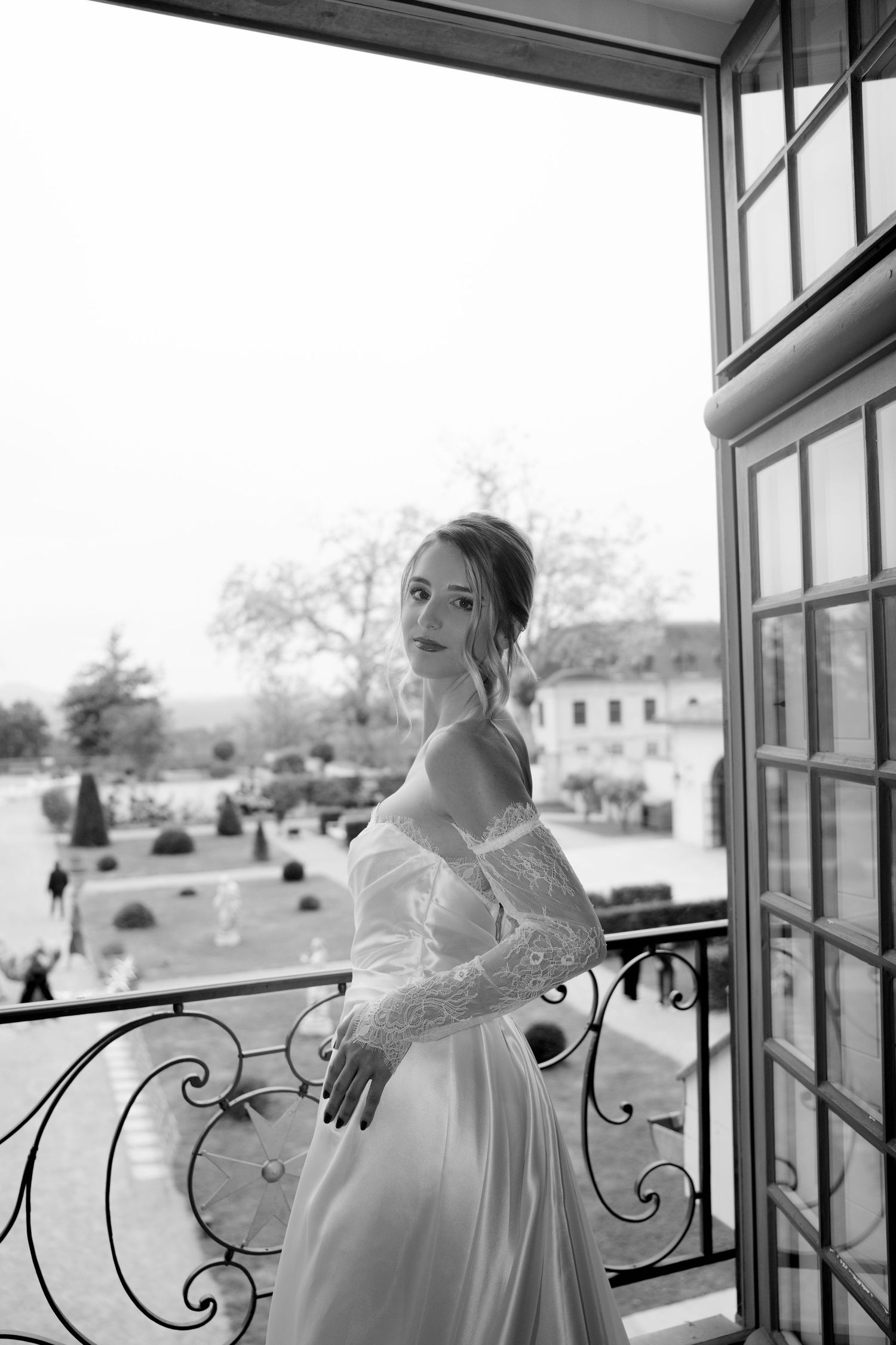 Black and white portrait of bride in lace wedding dress posing on balcony with garden view.