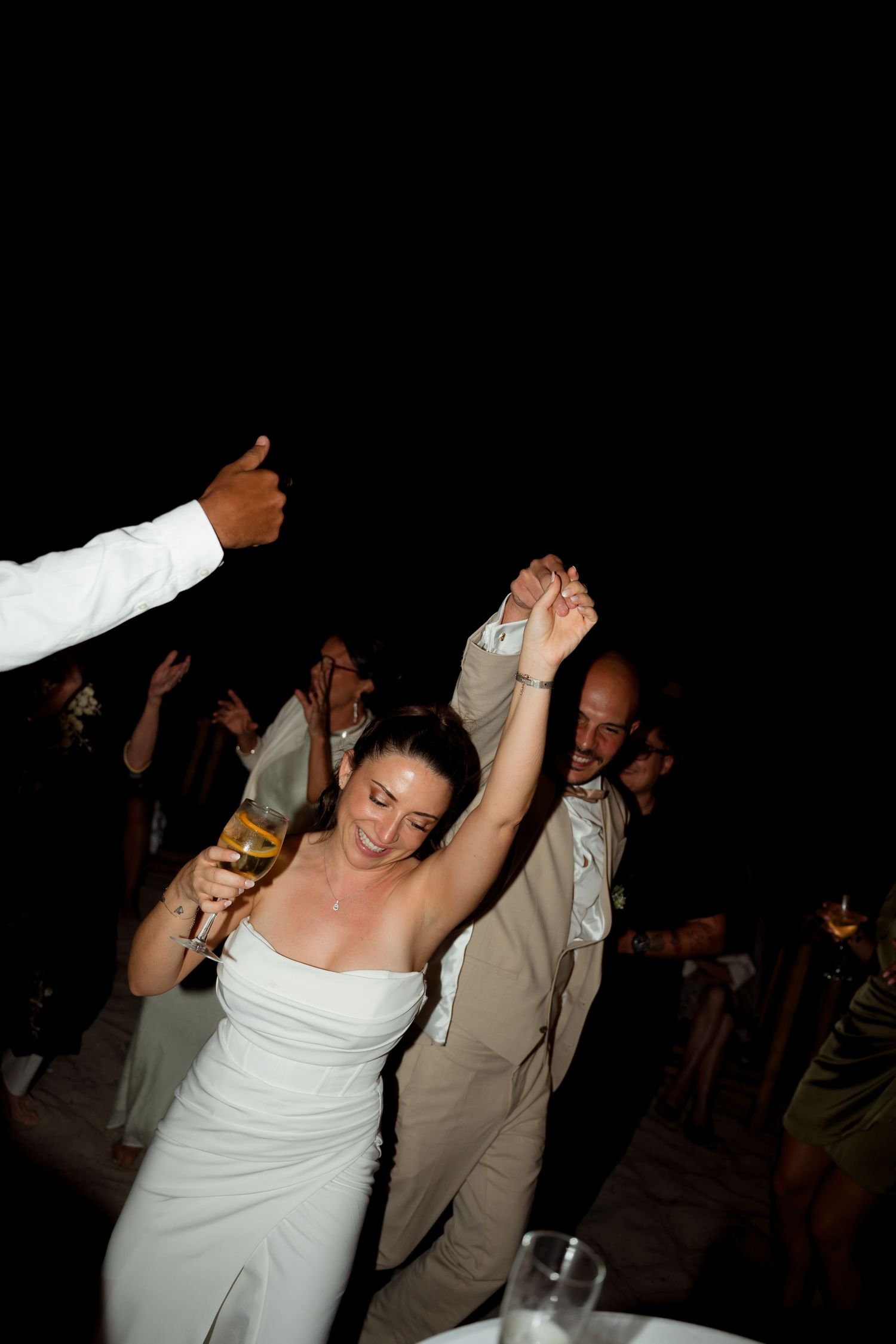 Bride in white strapless gown dancing joyfully with raised arms at evening wedding reception.