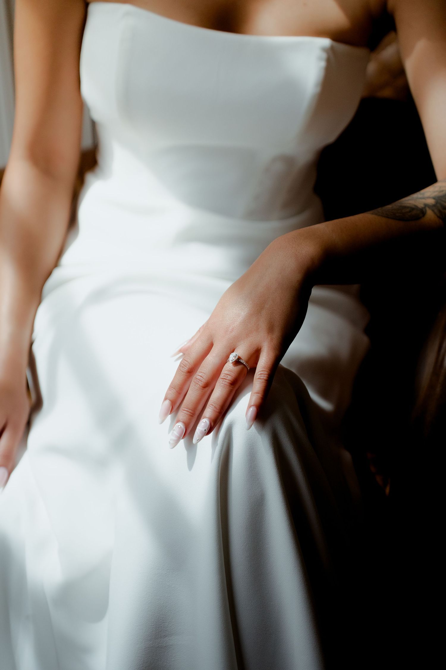 Close-up of bride's hand with long nude nails and gemstone ring resting on white satin wedding gown.