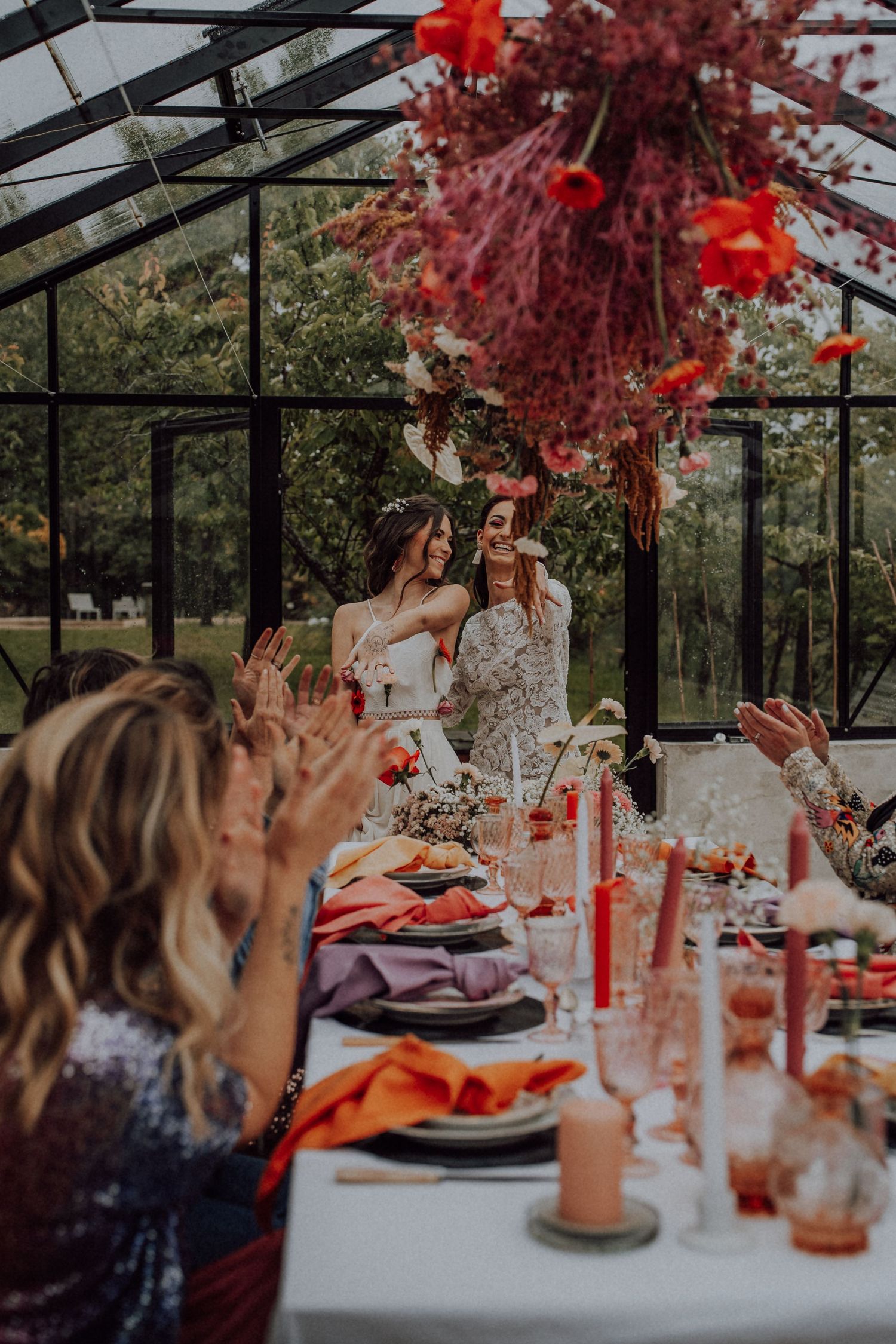 Two brides sharing a joyful moment at a boho wedding reception in a greenhouse with vibrant floral decor.