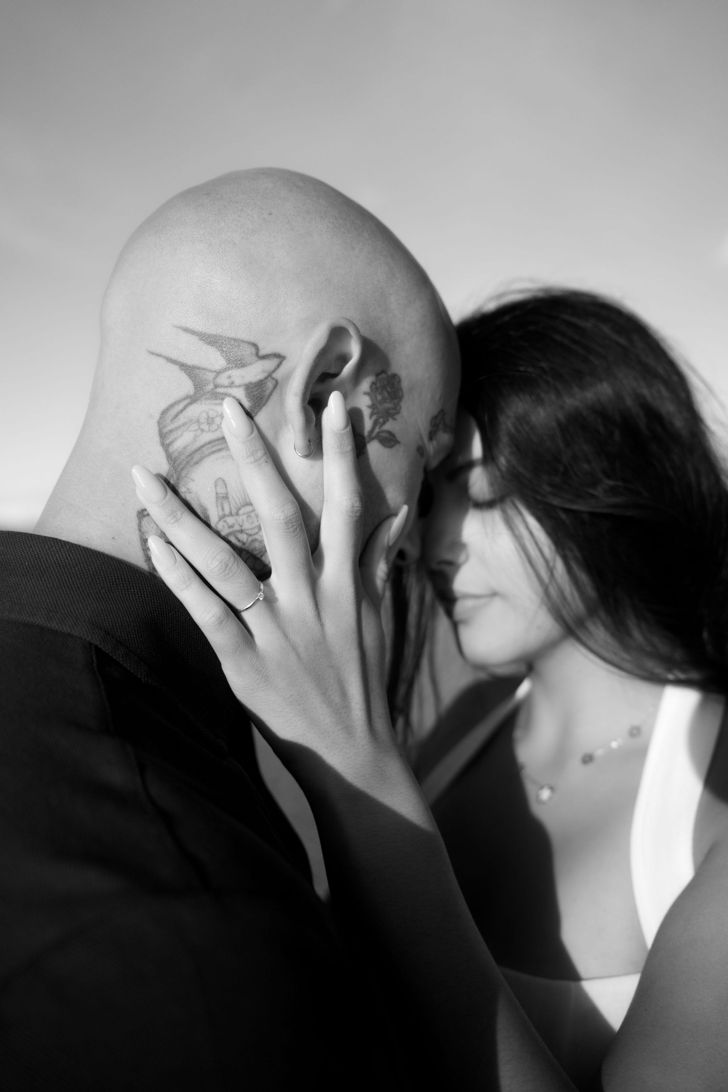 Black and white close-up of couple embracing, woman's hand with ring resting on man's tattooed head.