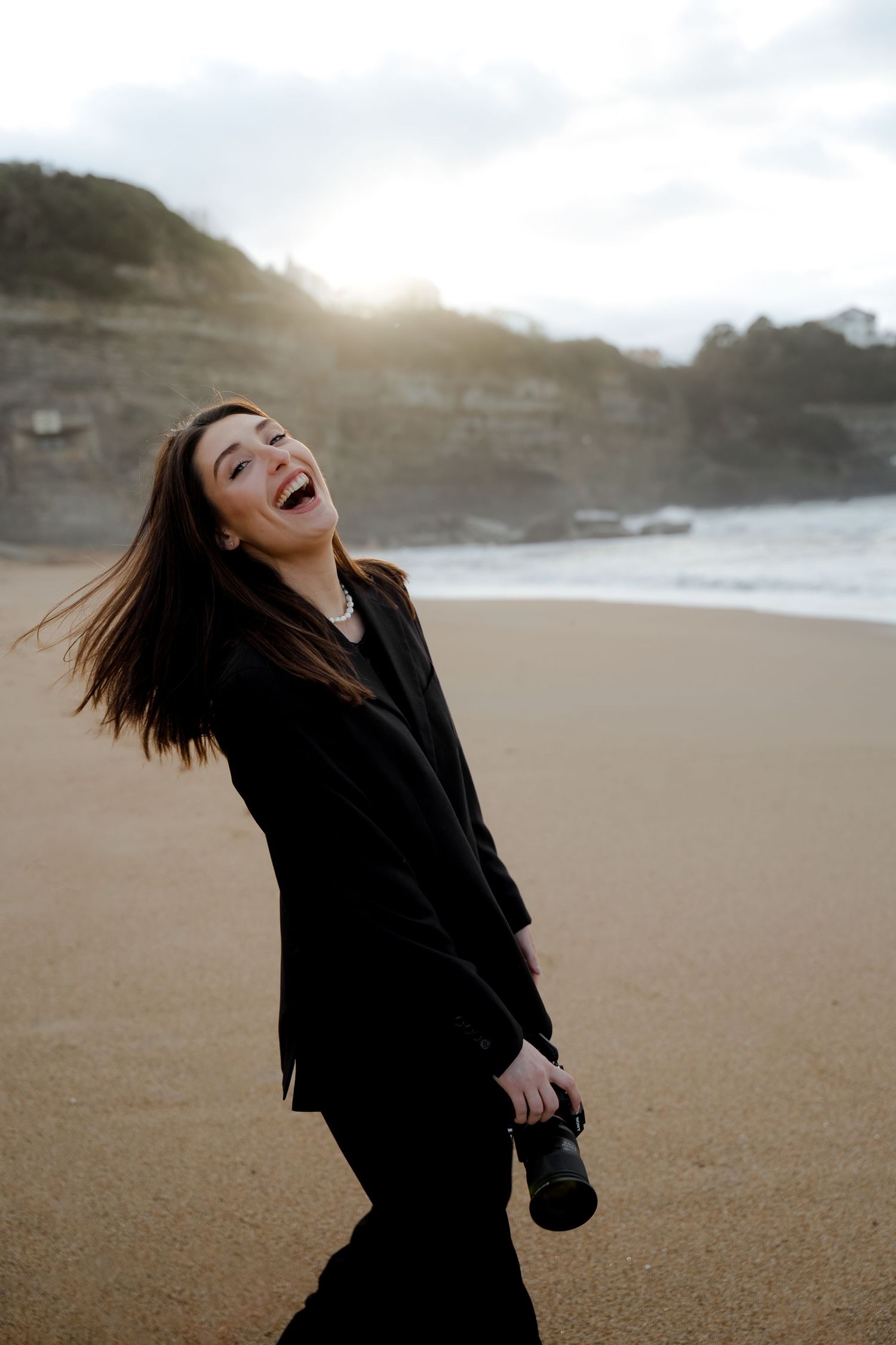 Joyful woman in black outfit laughing on beach holding camera, hair blowing in wind.