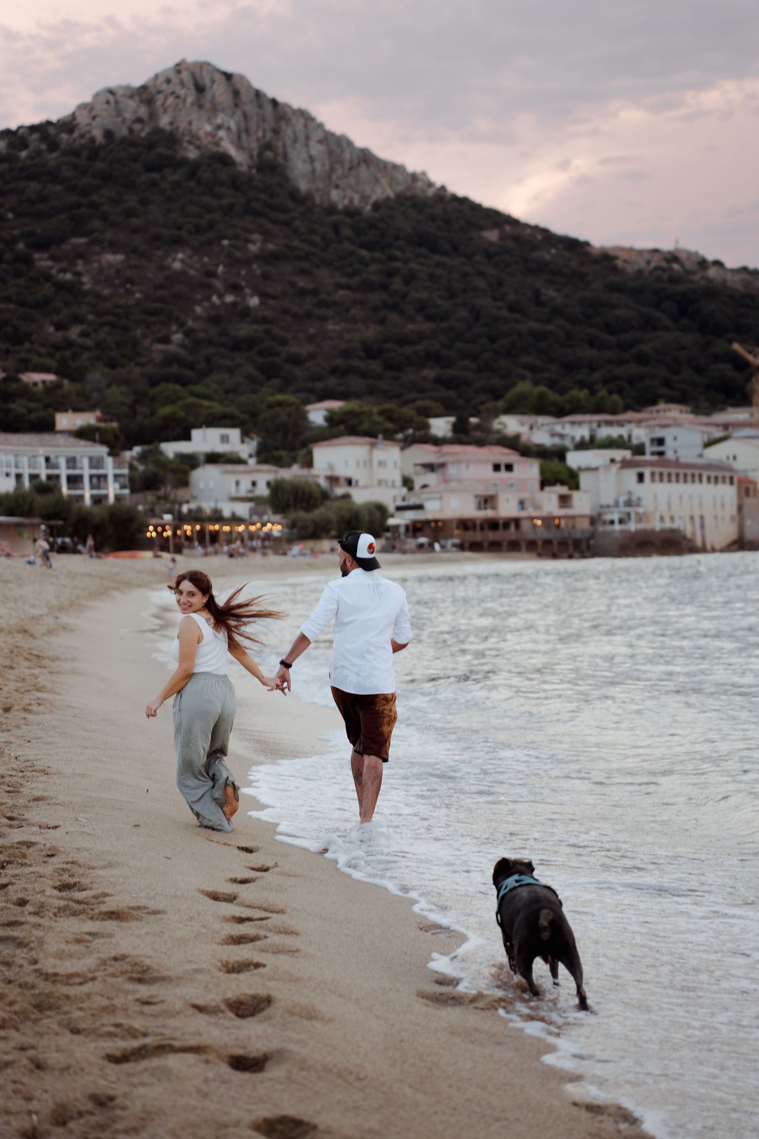 Couple running hand in hand along a sandy beach at dusk with their black dog and mountain backdrop.