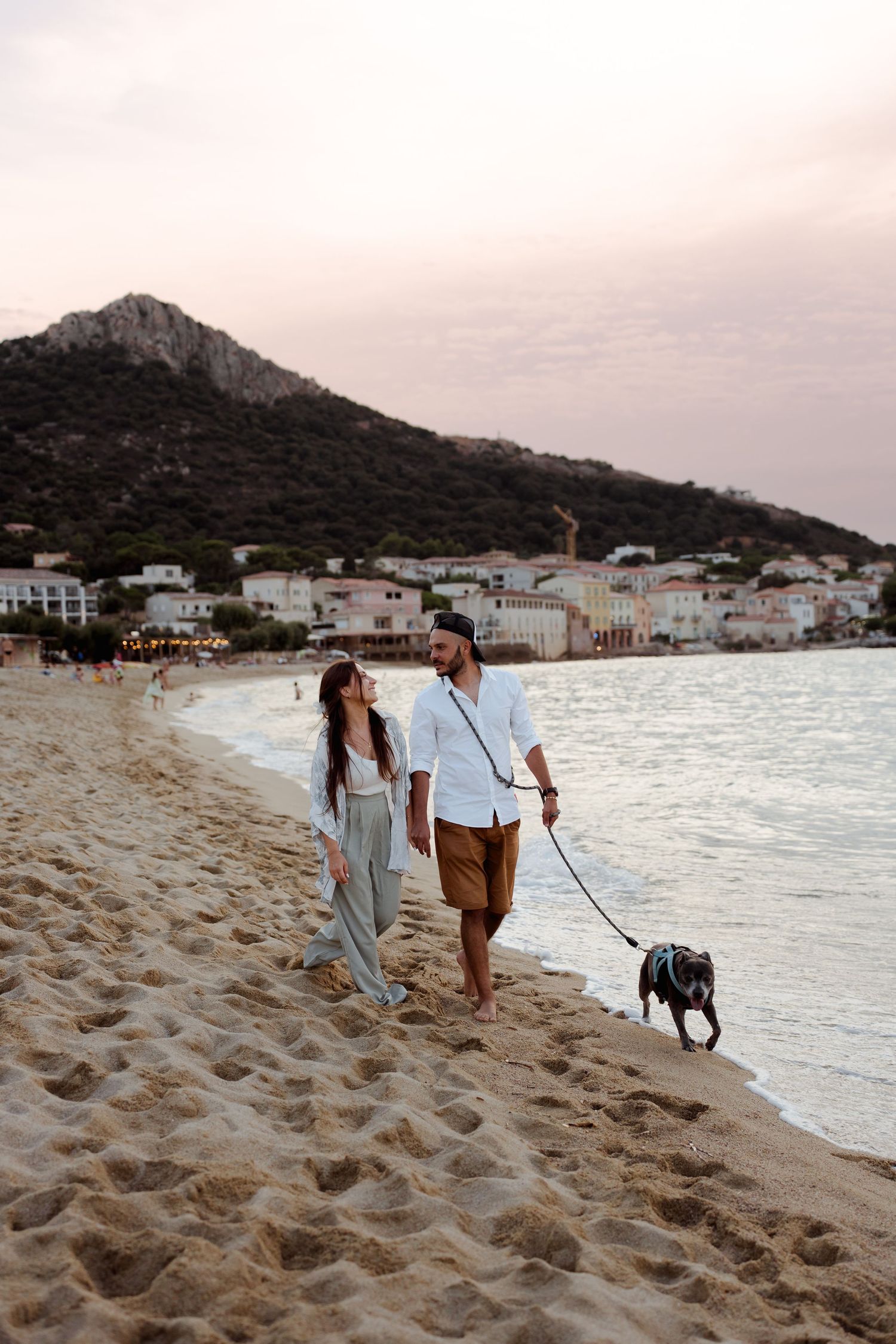 Couple walking their black dog along a sandy beach at sunset with a mountain village behind them.