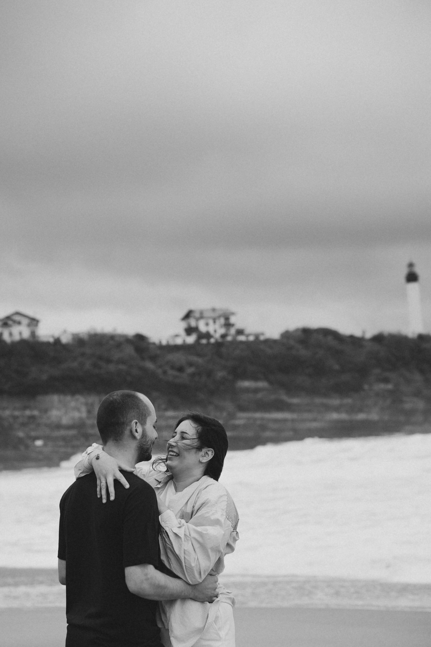 Black and white photo of a laughing couple embracing on a beach with a lighthouse behind them.