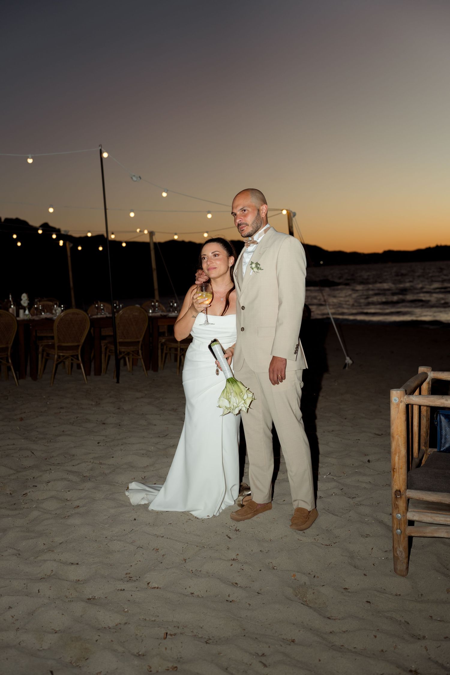 Bride and groom pose romantically on sandy beach at twilight with string lights and reception tables.