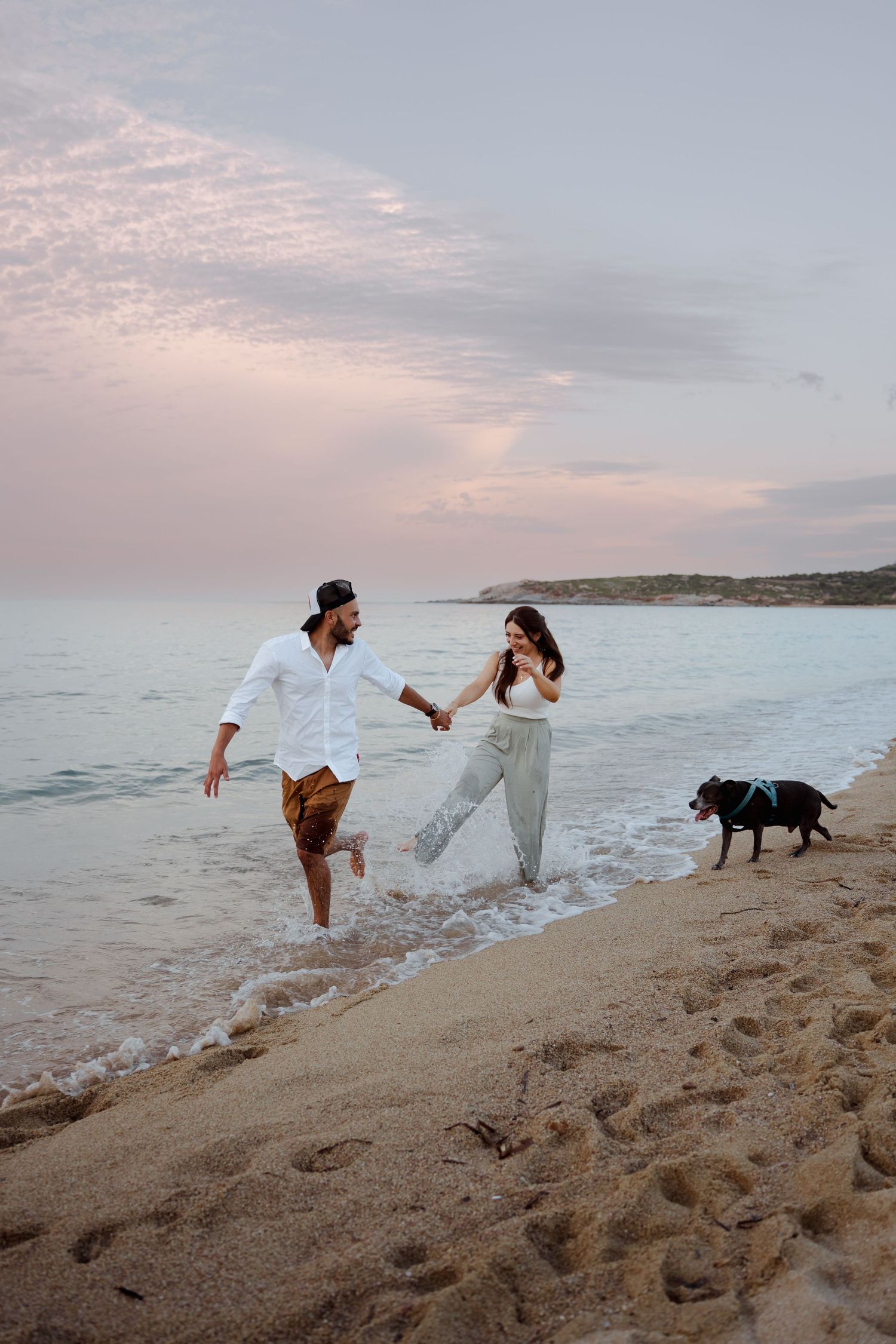 Happy couple holding hands and playing in ocean waves on a sandy beach at sunset with their black dog.