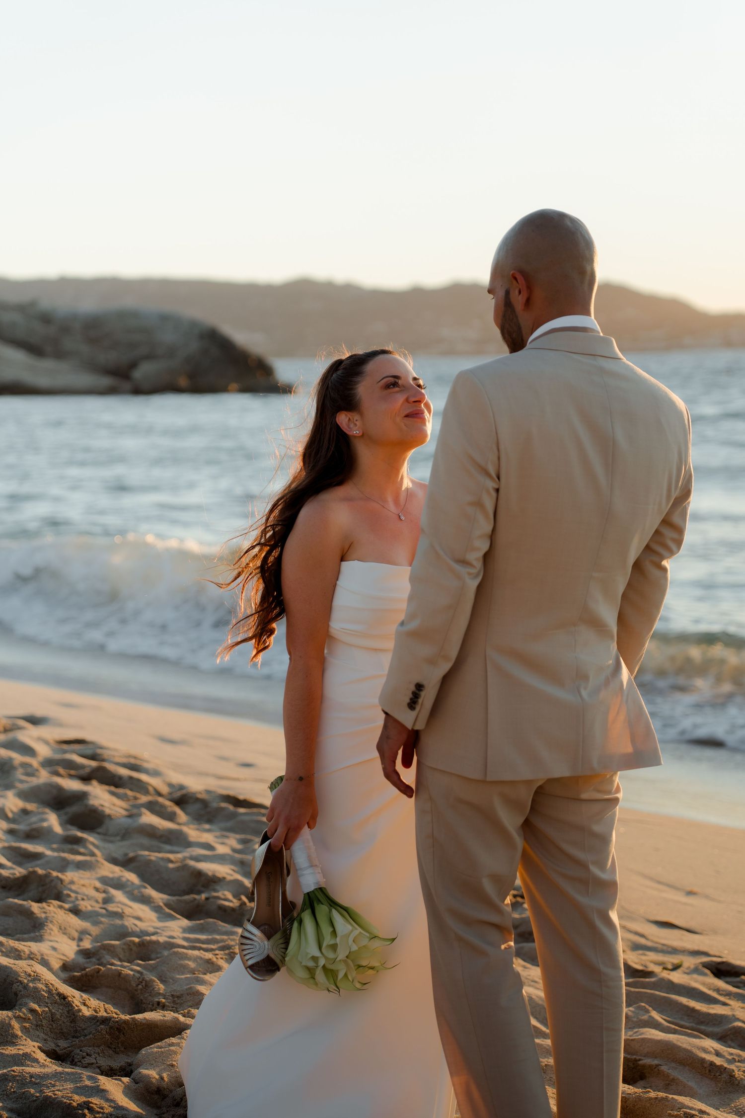 Smiling bride holding bouquet looks back at groom on sandy beach at golden sunset.