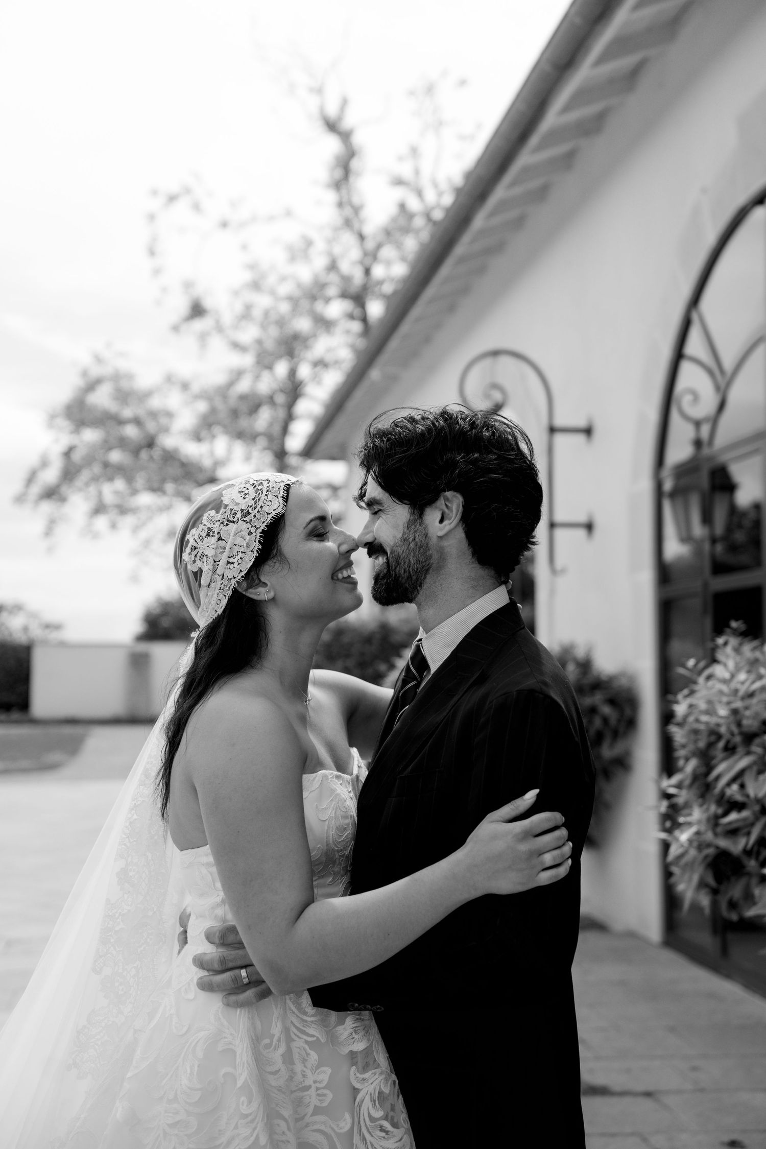 Black and white close-up of bride in lace headpiece and bearded groom sharing a tender moment outside.