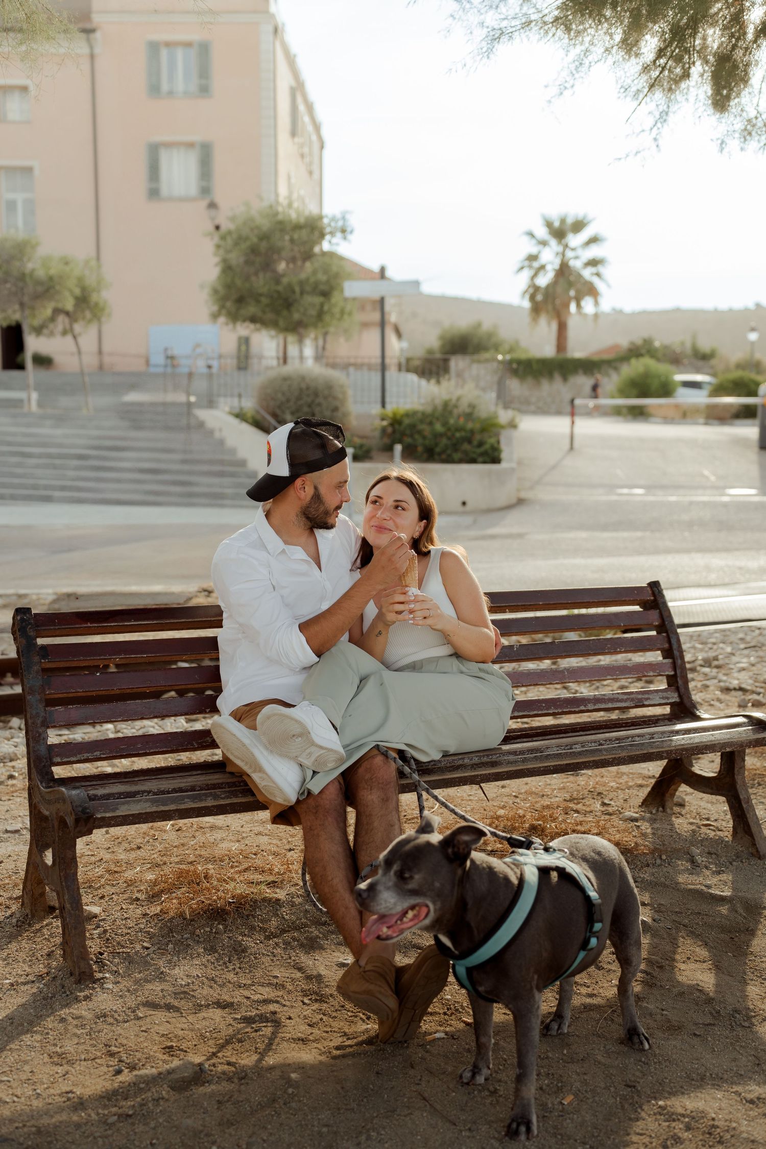 Happy couple sitting on a wooden bench with their gray dog on a leash in a sunny town square.