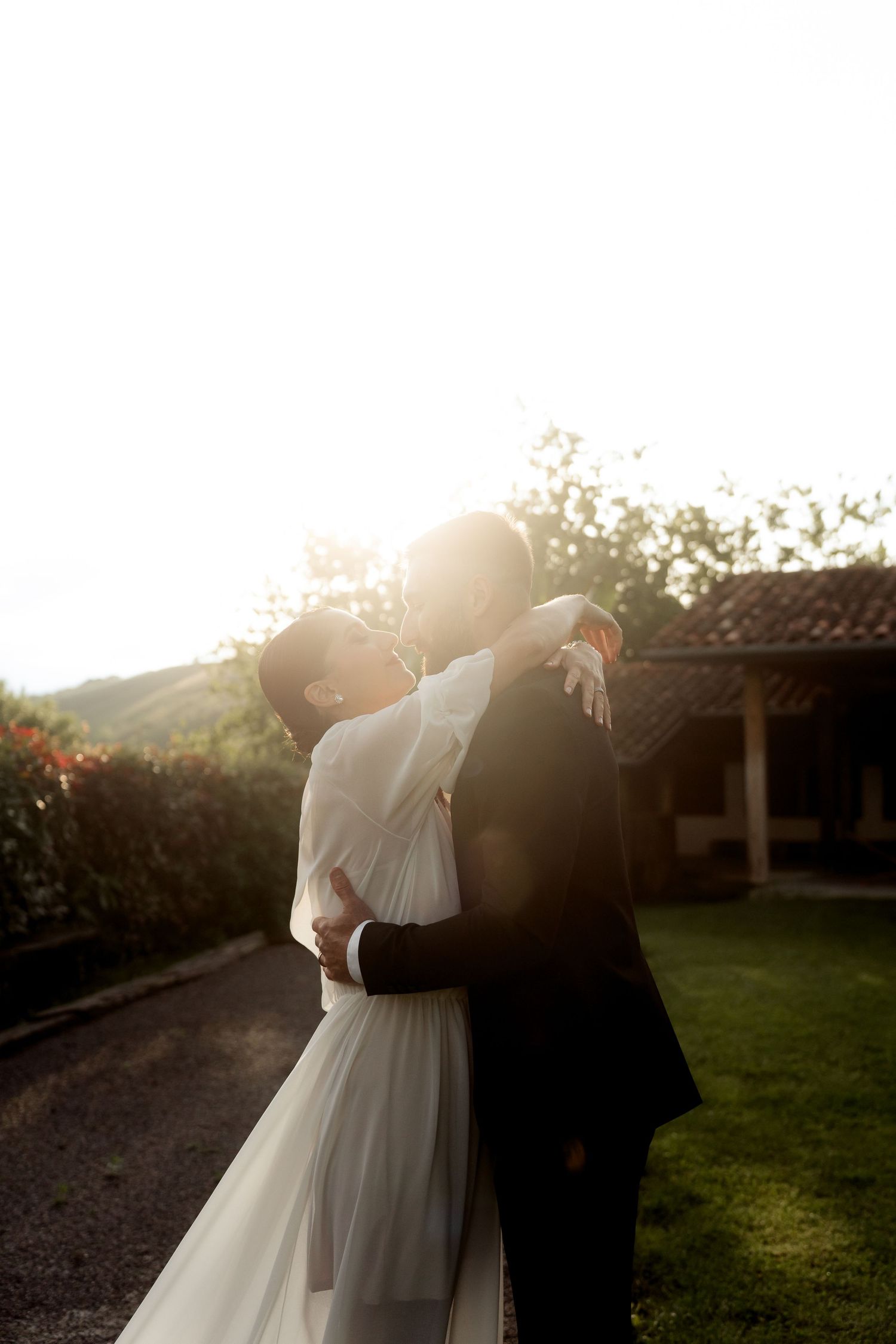 Bride and groom embrace romantically in glowing golden hour sunlight in garden setting.