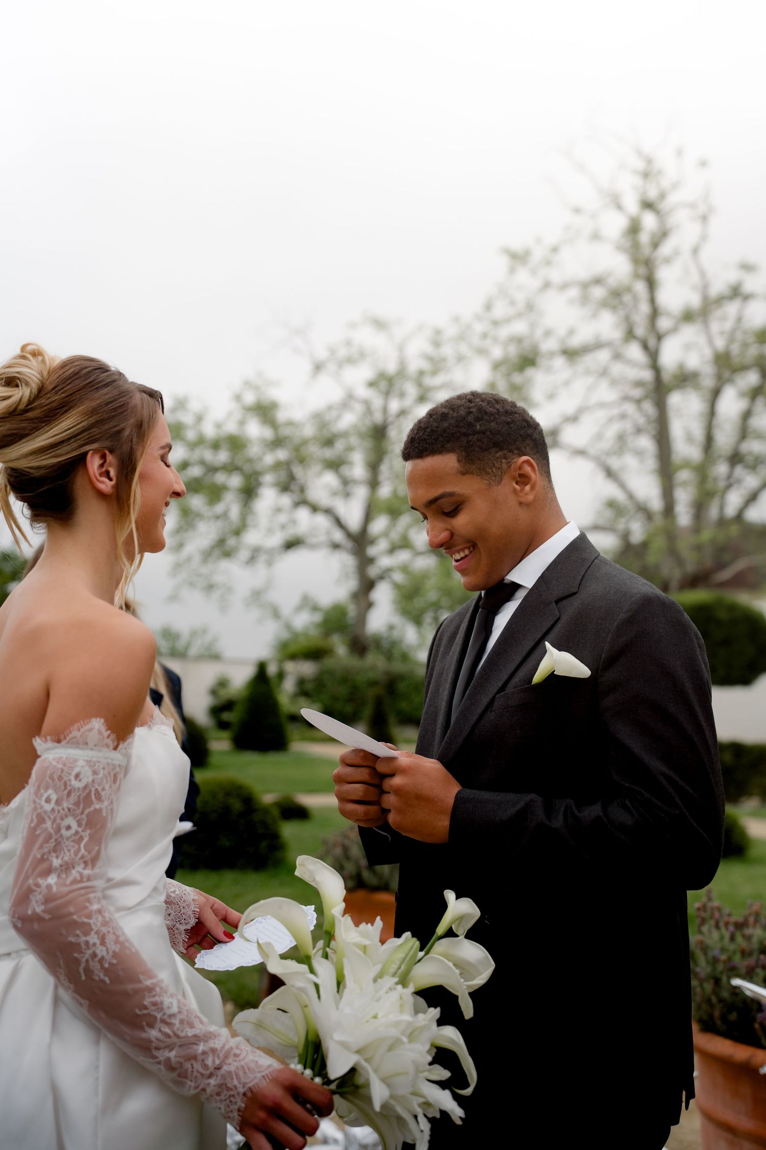 Groom in black suit reading wedding vows to smiling bride holding white lily bouquet outdoors.