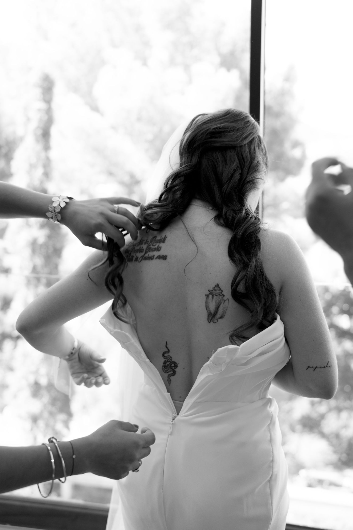 Black and white photo of bride with floral back tattoos having wedding dress fastened by helpers.